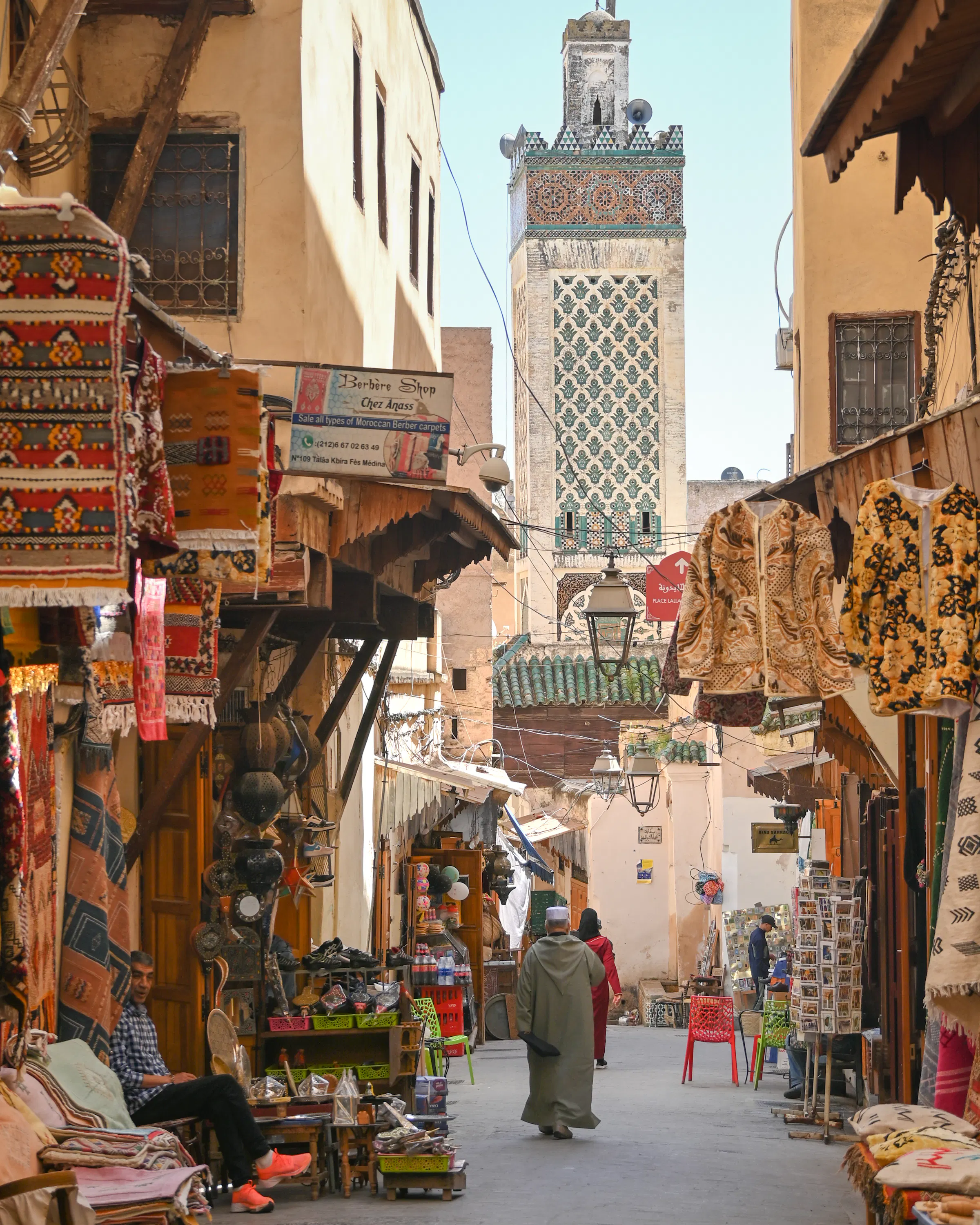 Narrow street lined with shops and pedestrians, with a tall tower in the distance.