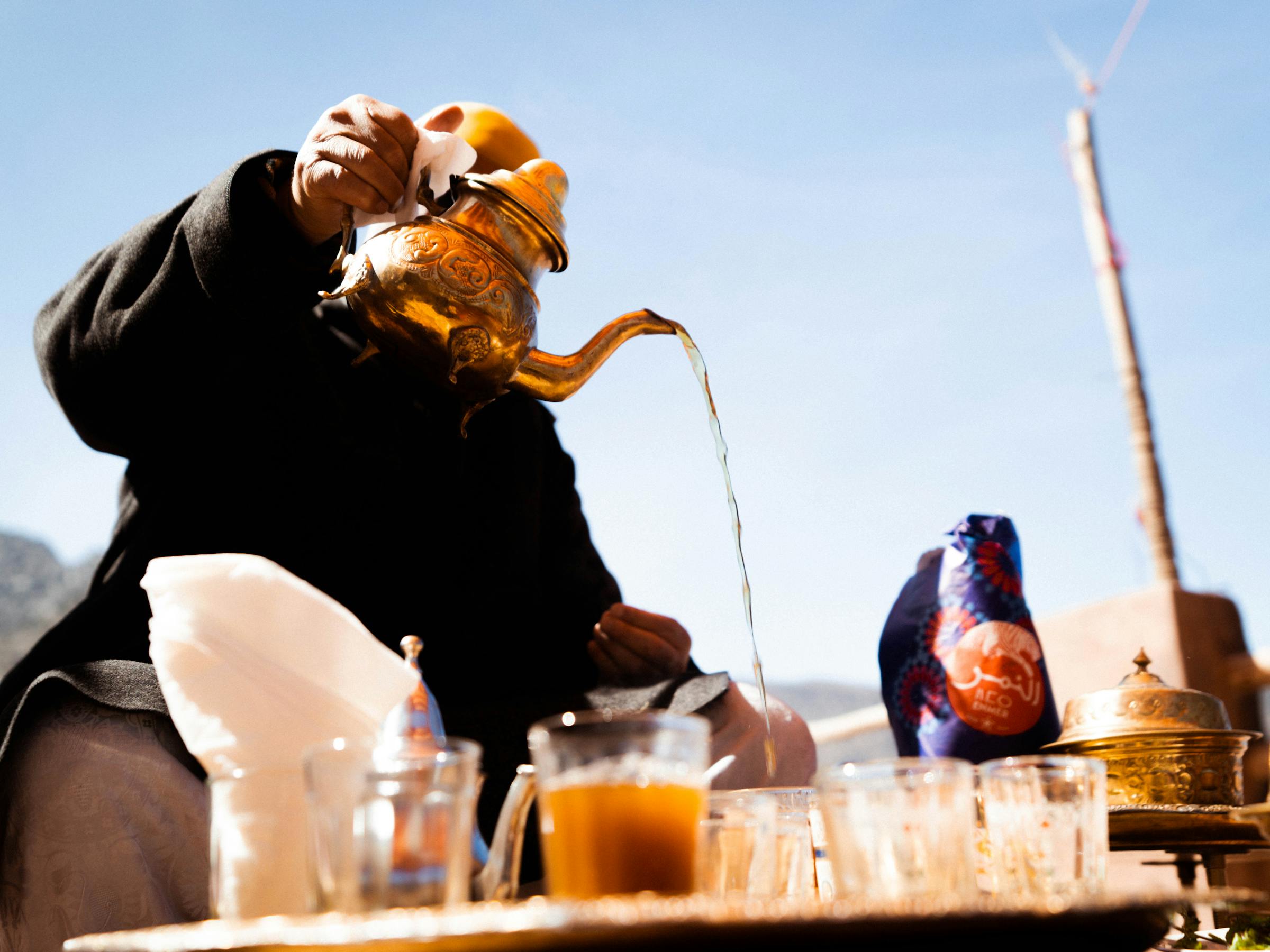 Person pouring tea from a teapot into small glasses on a sunlit table.