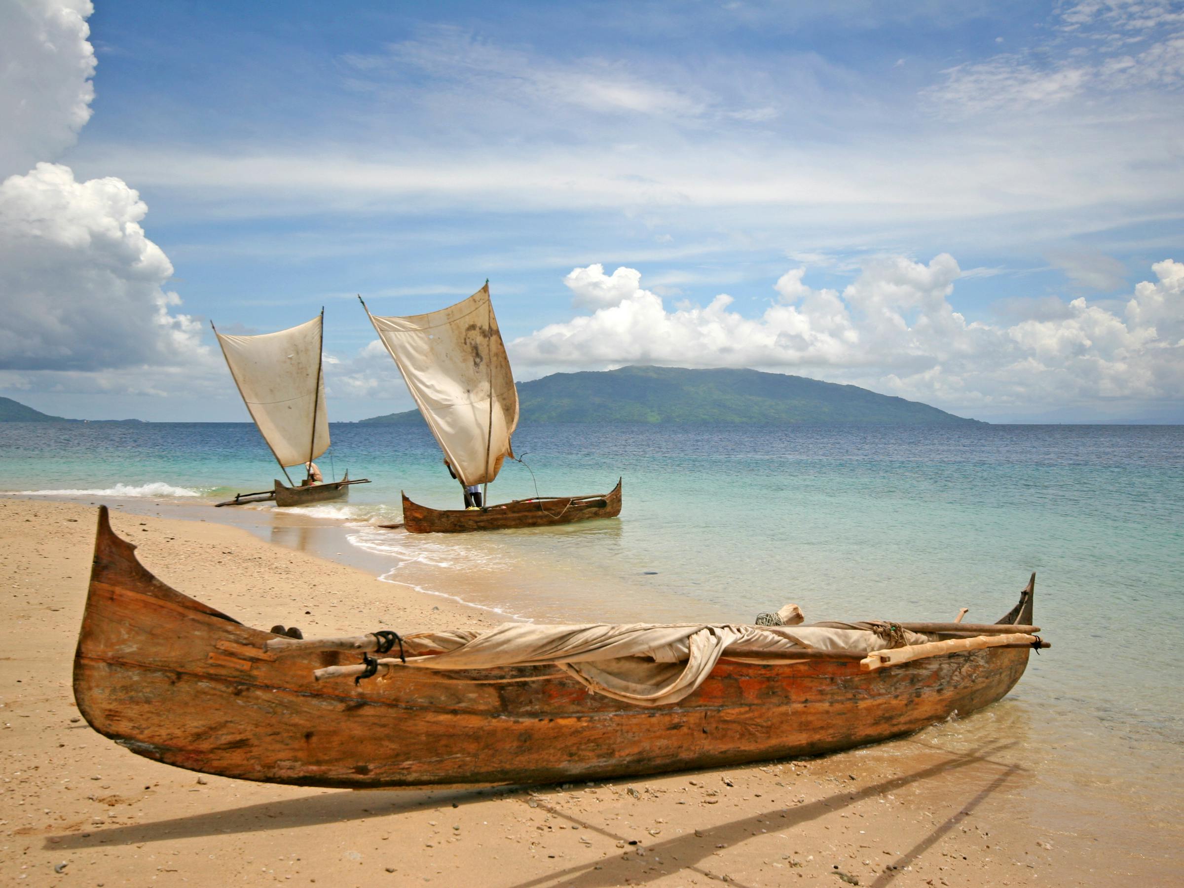 Weathered wooden canoe rests on a sandy shore as two small sailboats drift in calm, clear water under a blue sky.