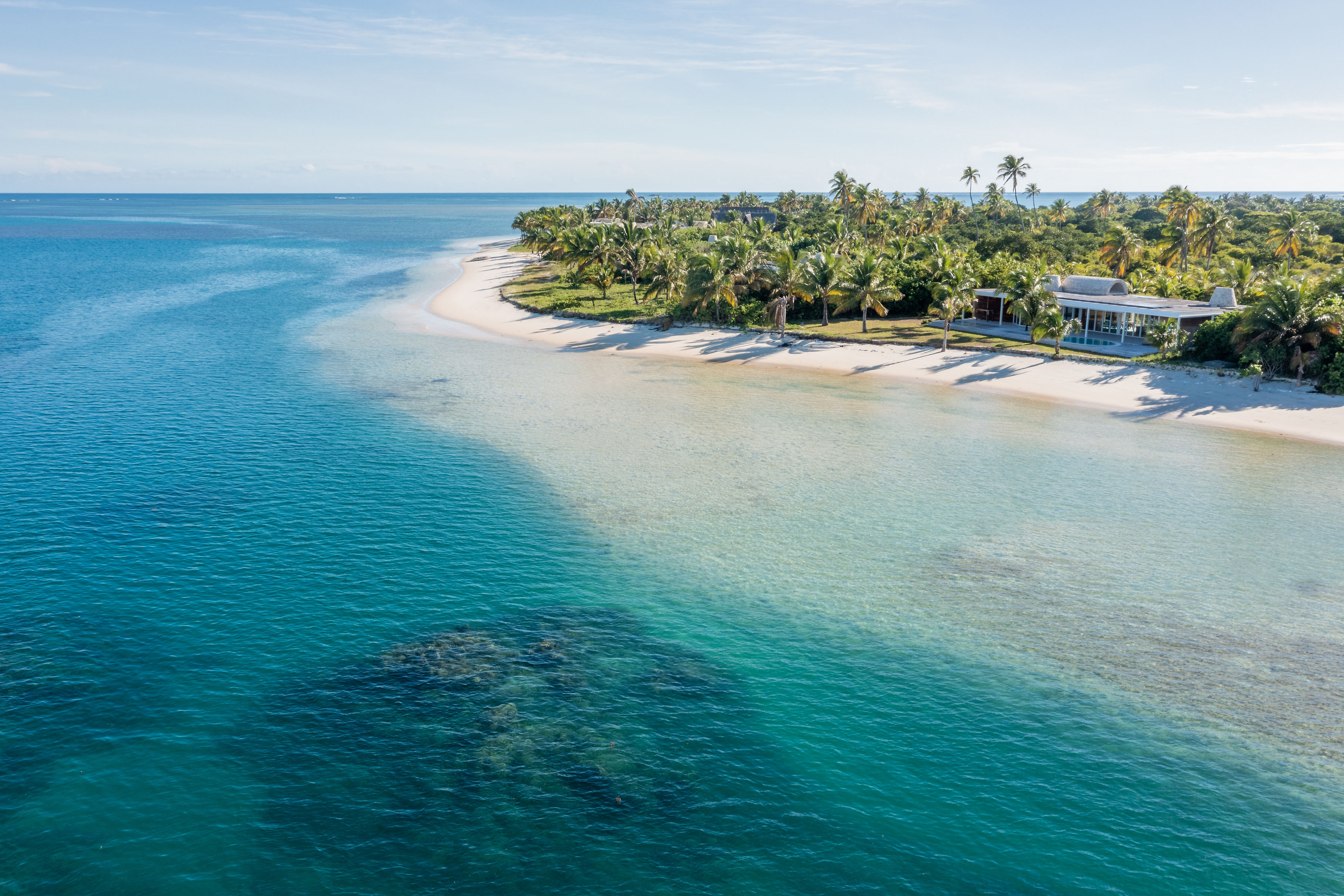 Aerial view of a palm-lined beach and resort buildings beside clear turquoise shallows fading into deep blue water.