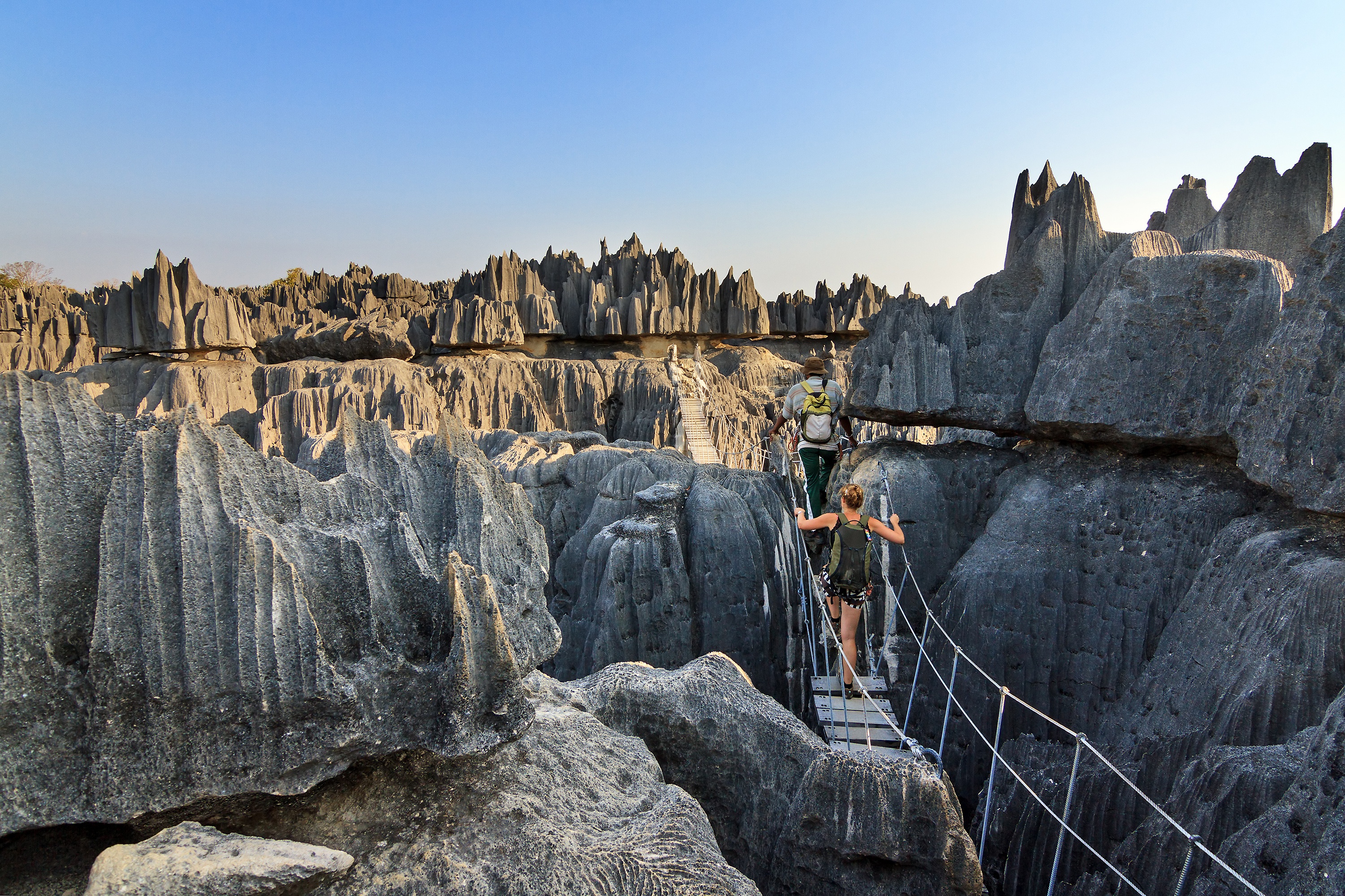 Two hikers cross a narrow suspension bridge through sharp gray limestone pinnacles under a clear blue sky.
