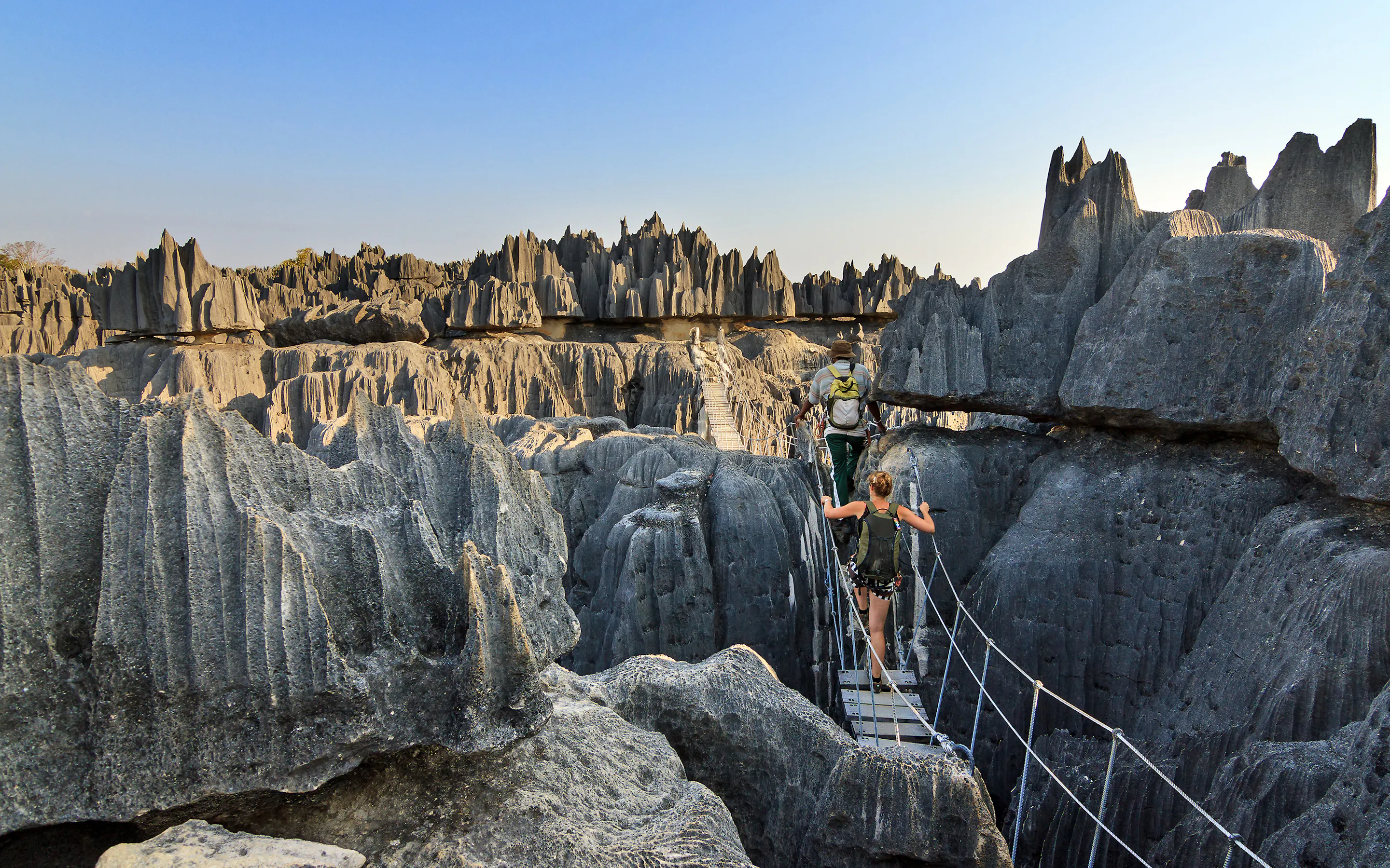 Two hikers cross a narrow suspension bridge through sharp gray limestone pinnacles under a clear blue sky.