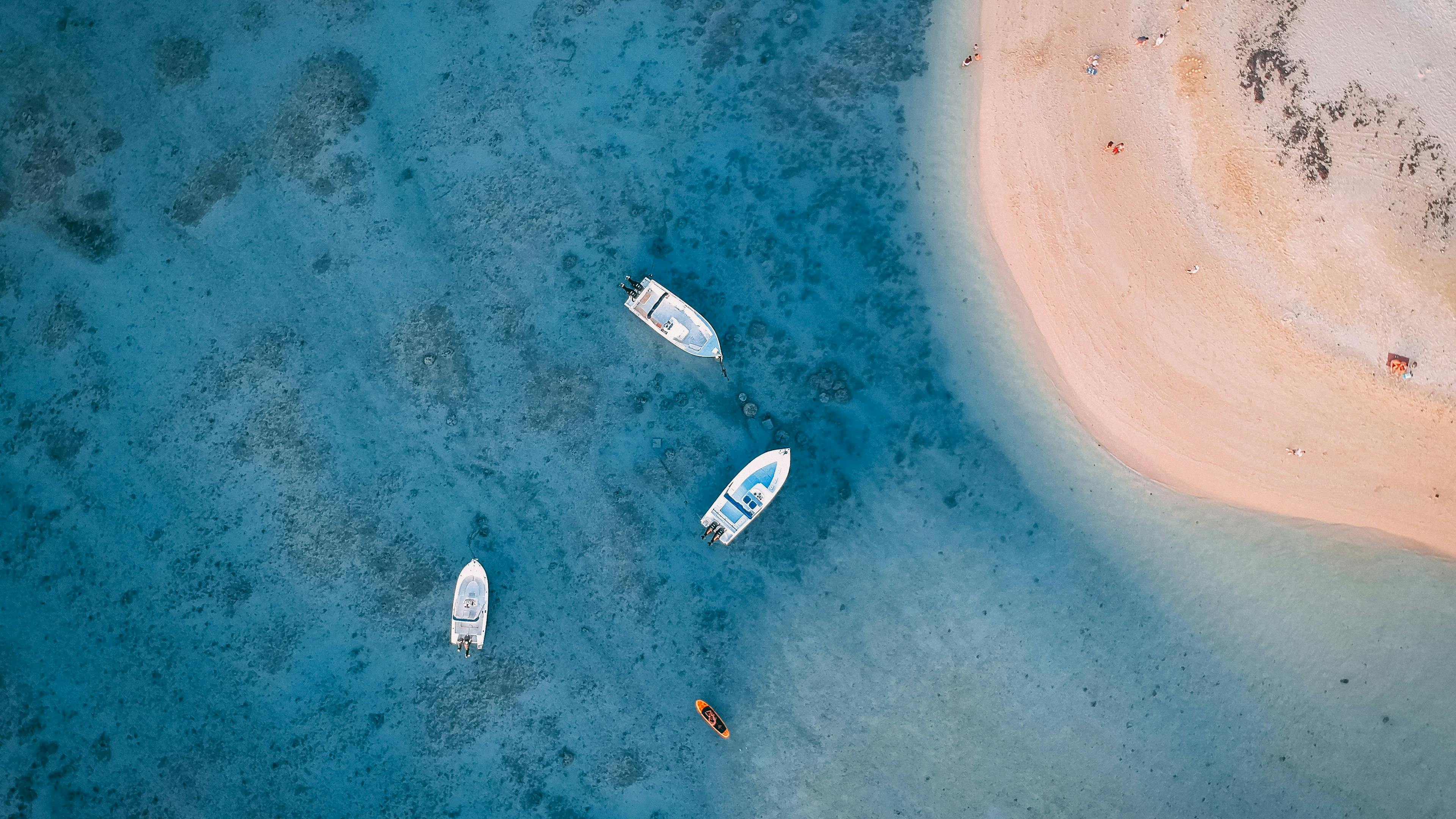 Aerial view of a pale sandbar in blue water, with two small boats anchored nearby and darker reef patches.