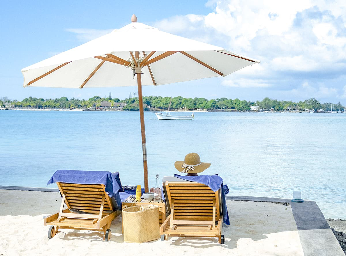 Two beach loungers and a large white umbrella face a calm bay, with a sailboat floating offshore under a bright sky.