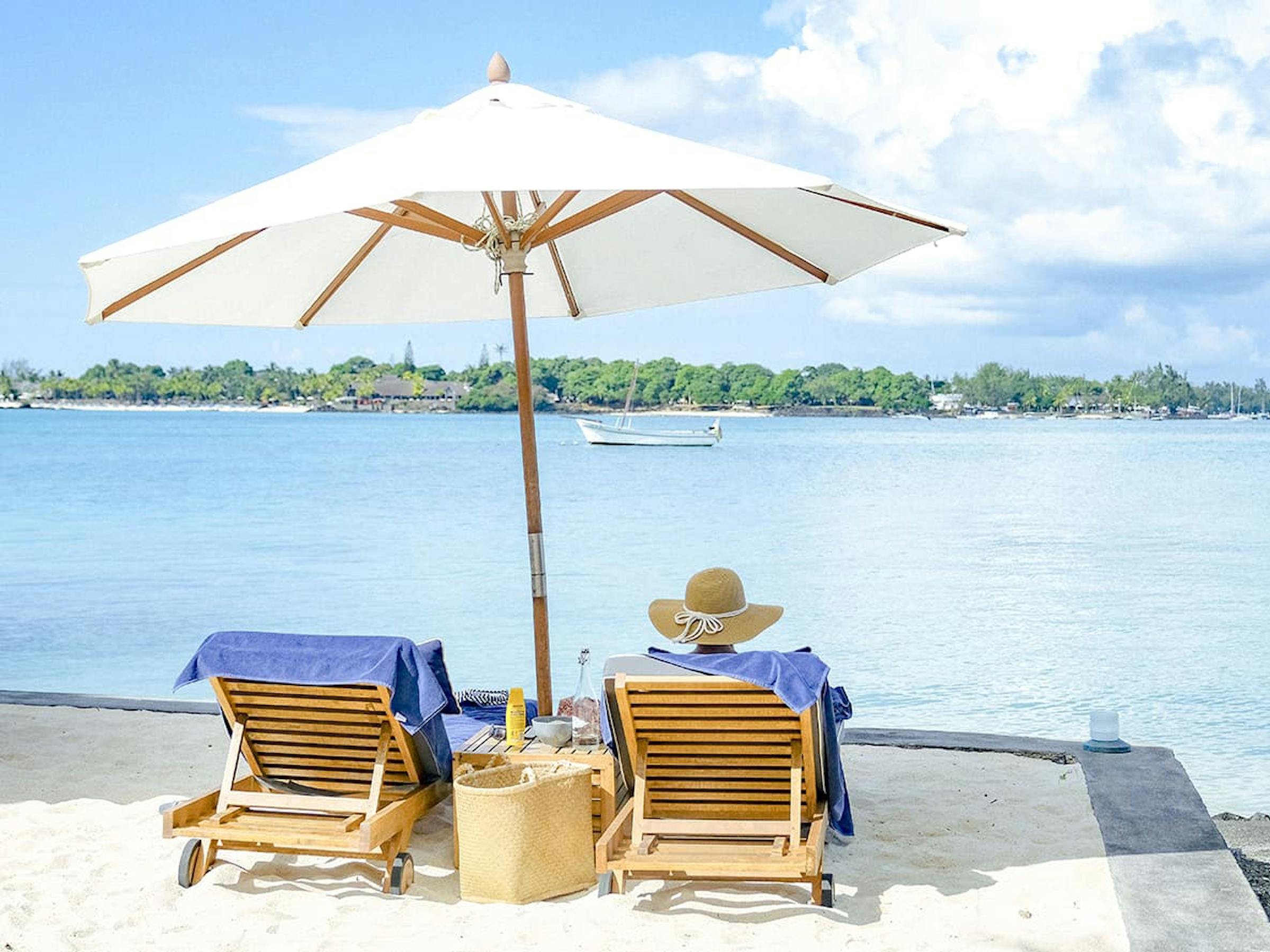 Two beach loungers and a large white umbrella face a calm bay, with a sailboat floating offshore under a bright sky.