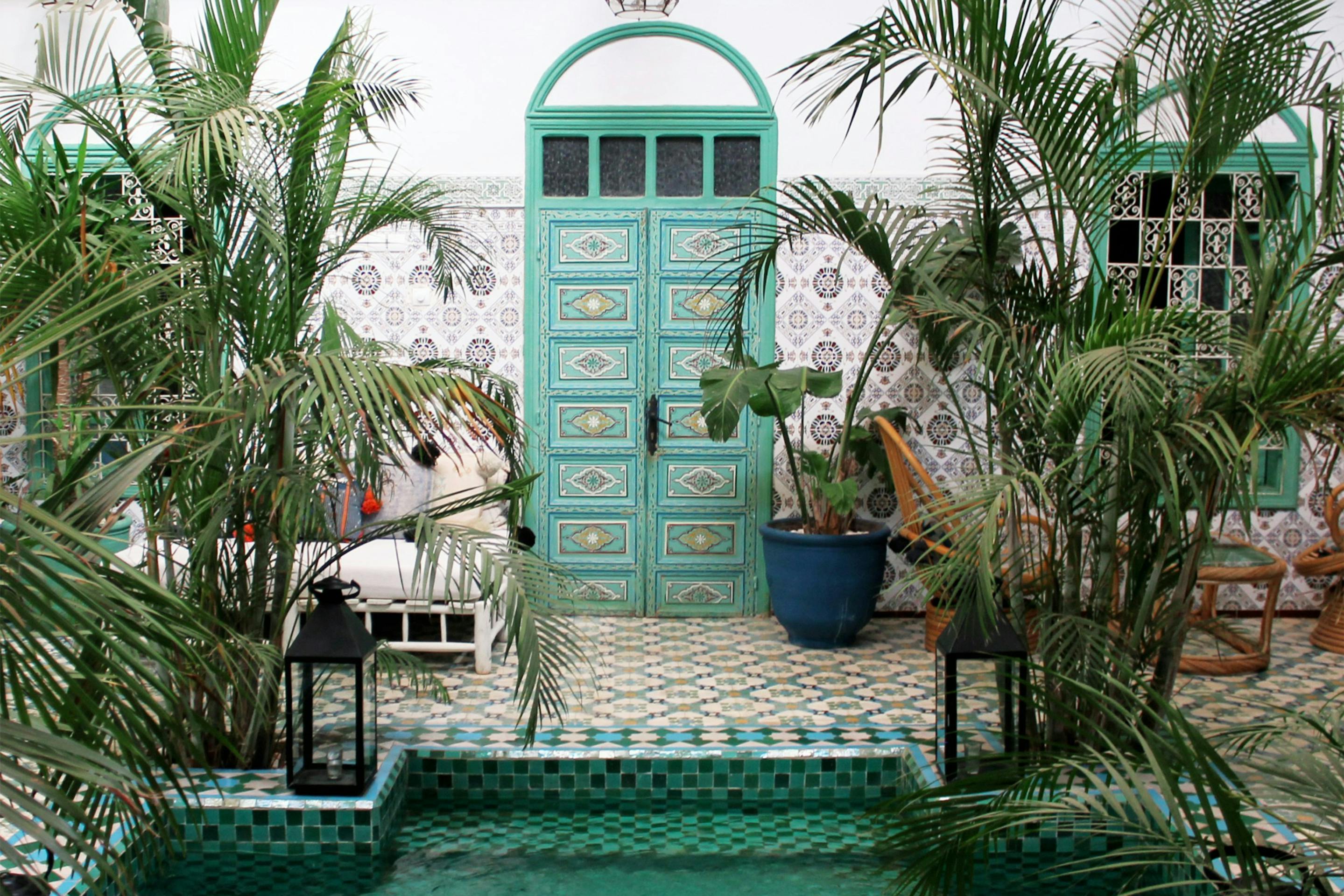 Turquoise door above a small pool surrounded by potted palms and patterned tile.