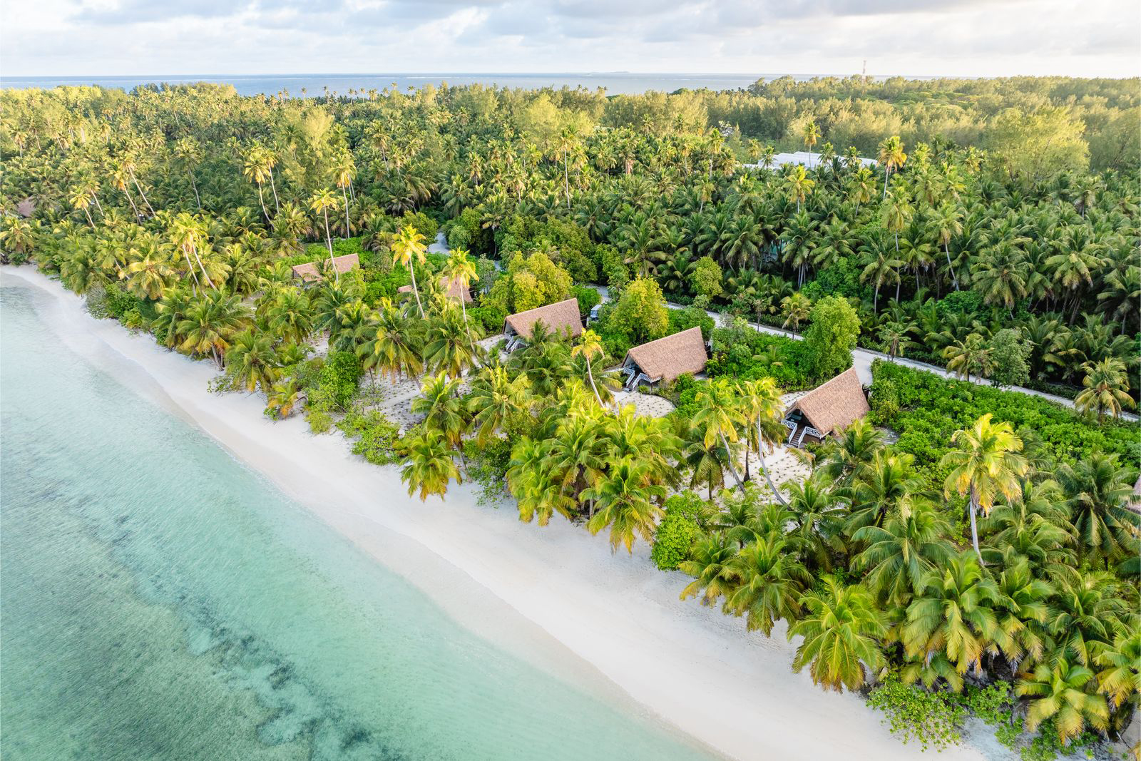 Aerial view of a white sand shoreline lined with palm trees and thatched huts beside clear turquoise water.