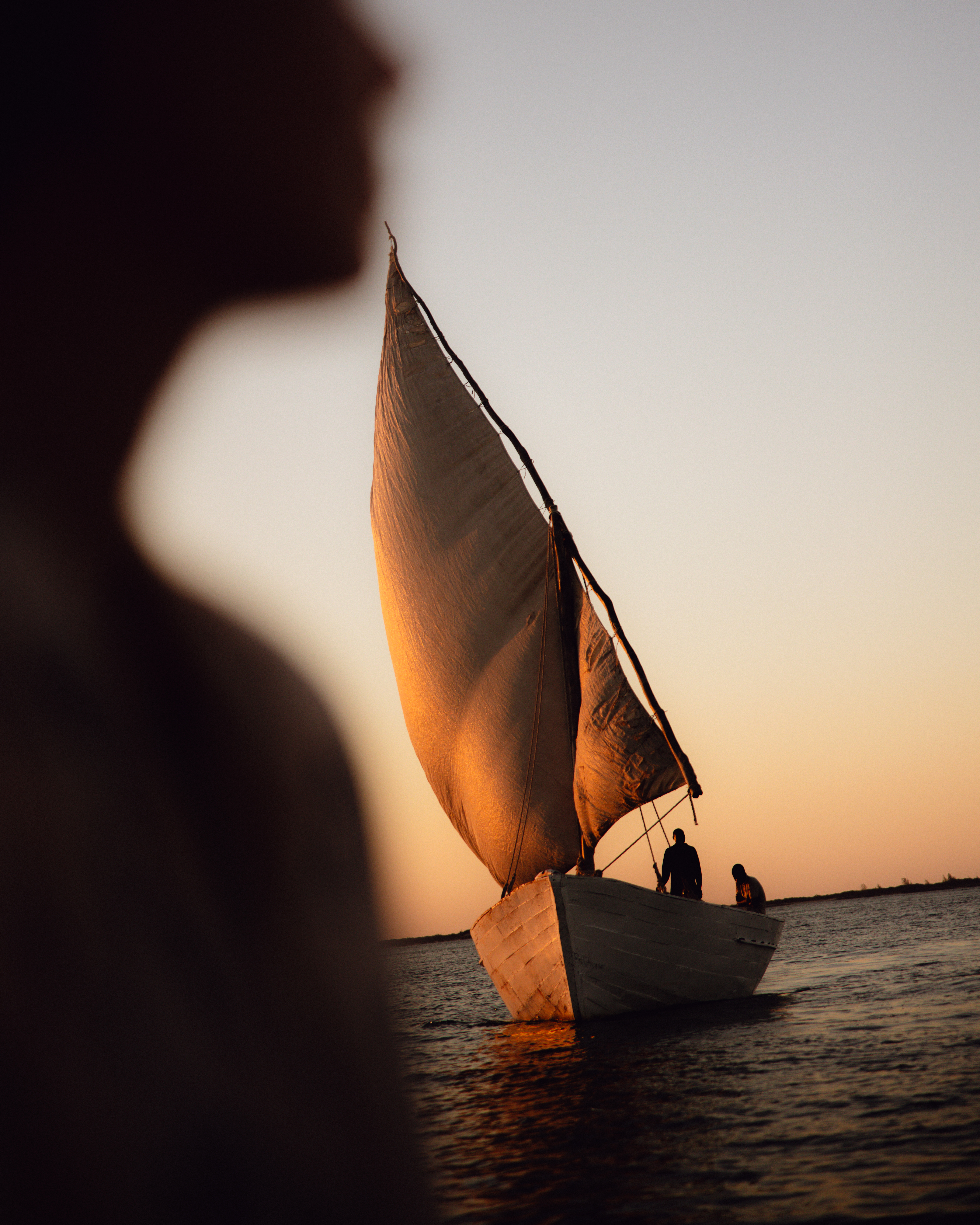 Small sailboat glides at sunset with a dark sail catching light, while a blurred figure watches from the foreground.