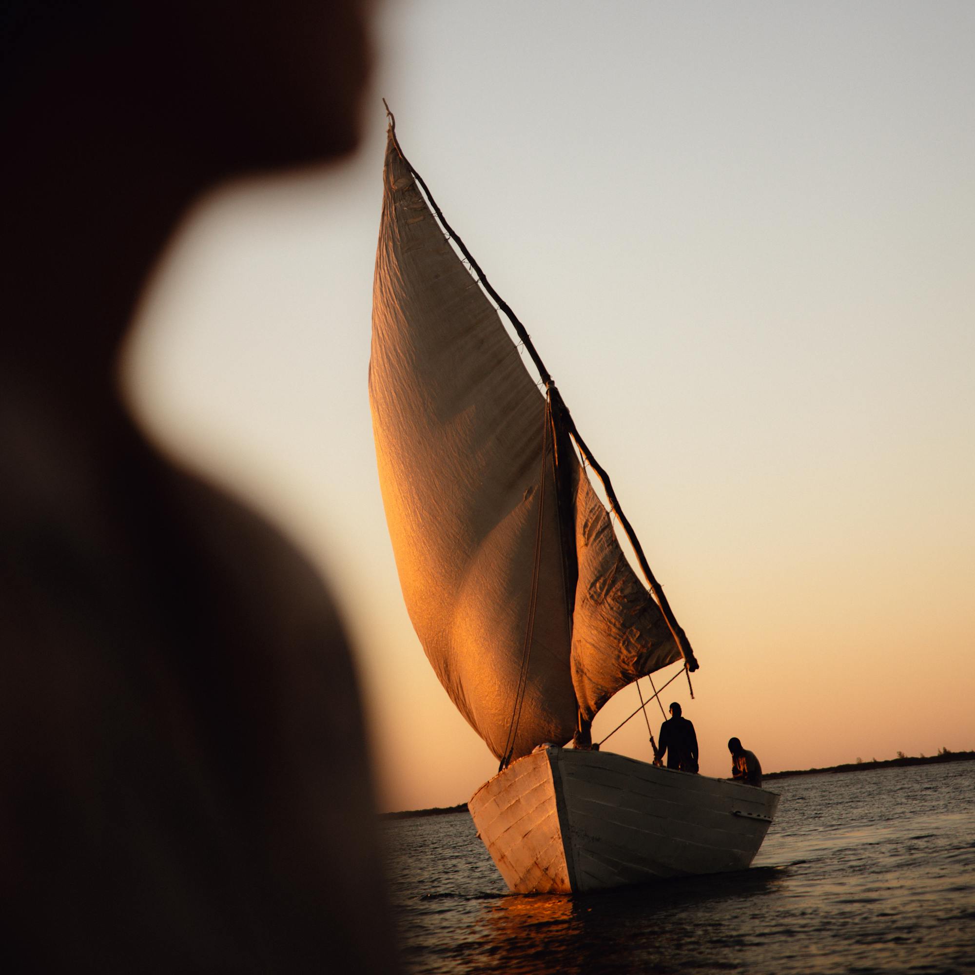 Small sailboat glides at sunset with a dark sail catching light, while a blurred figure watches from the foreground.
