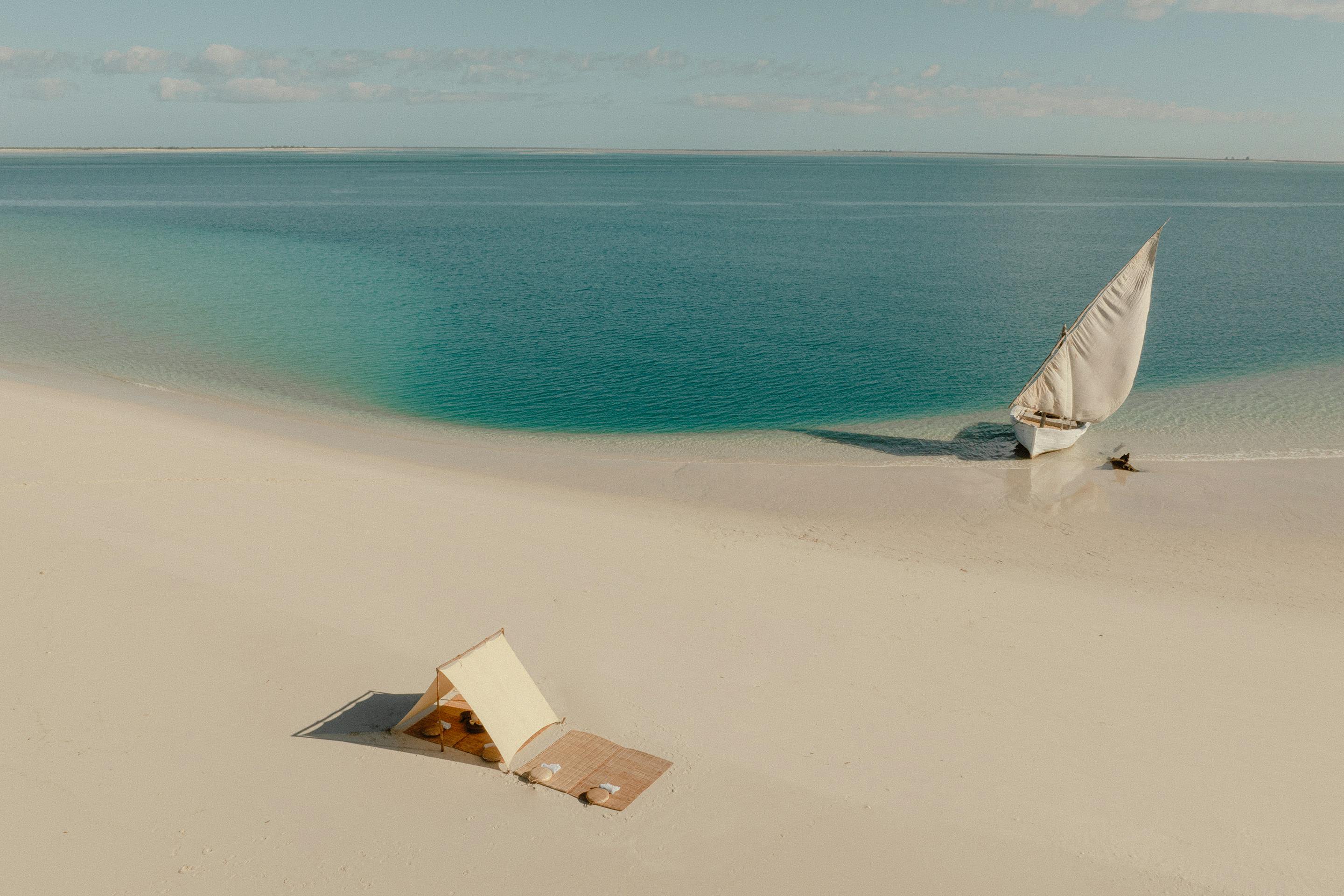 Aerial view of white sand and turquoise water with a small sailboat offshore and a shaded picnic setup below.