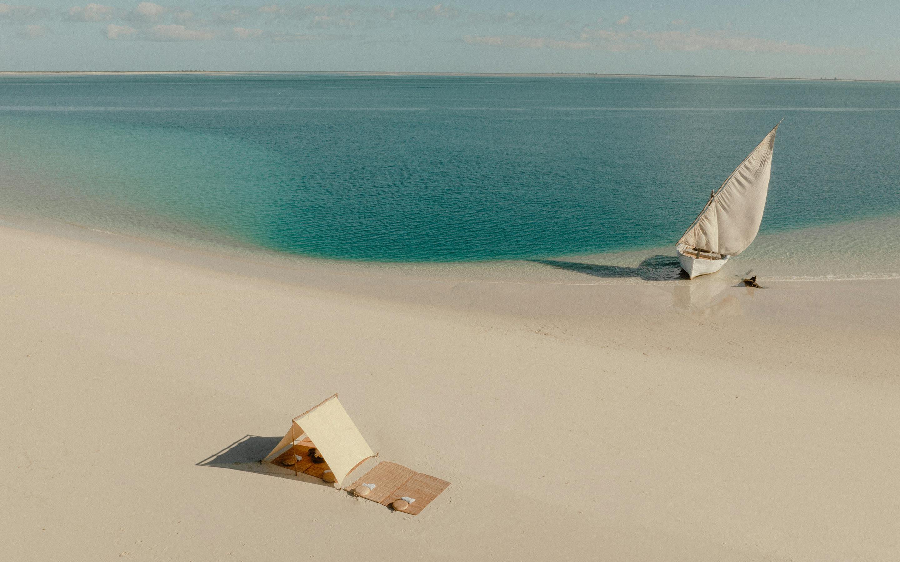 Aerial view of white sand and turquoise water with a small sailboat offshore and a shaded picnic setup below.