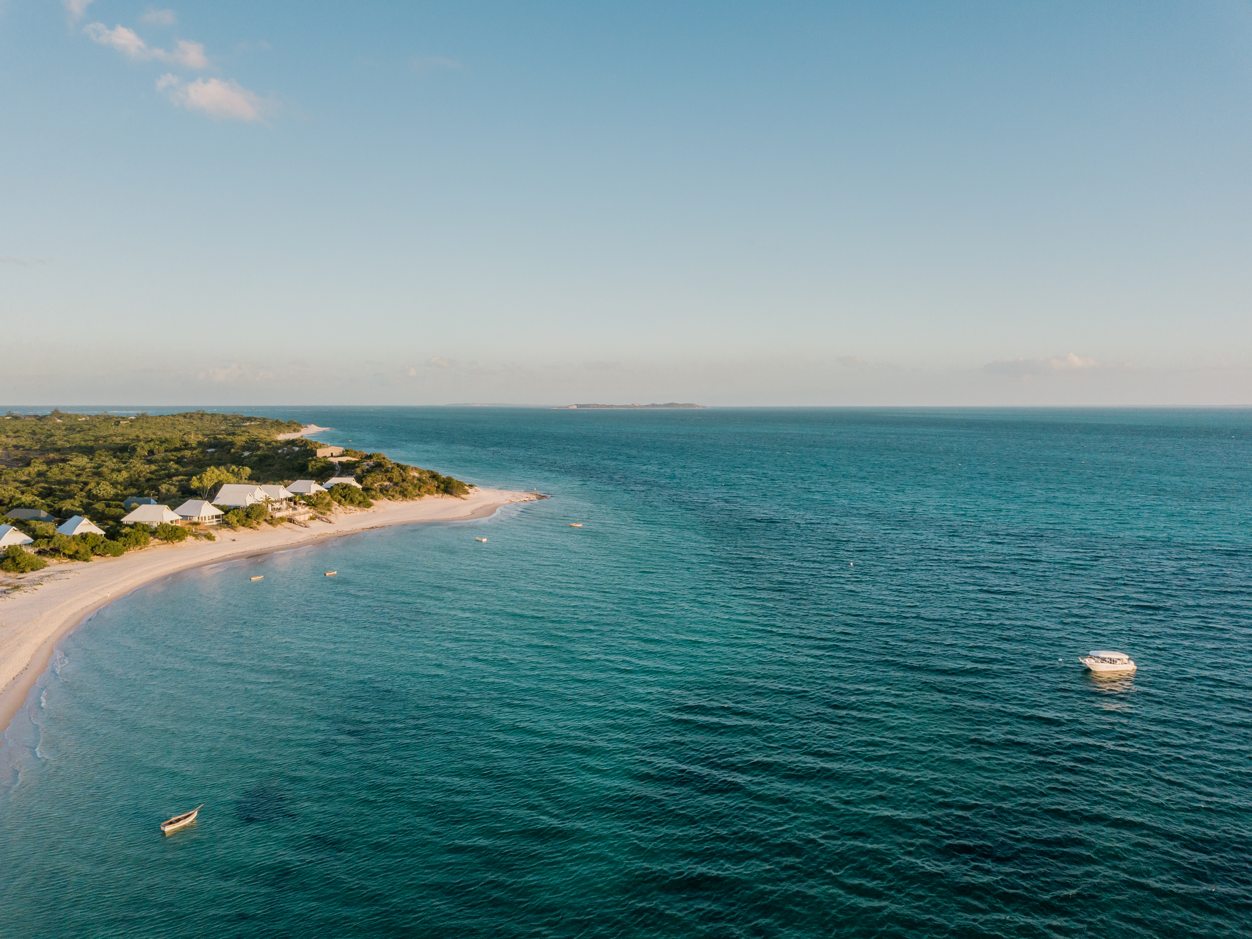 Aerial view of a long sandy shoreline and turquoise sea, with low buildings and trees lining the coast below.