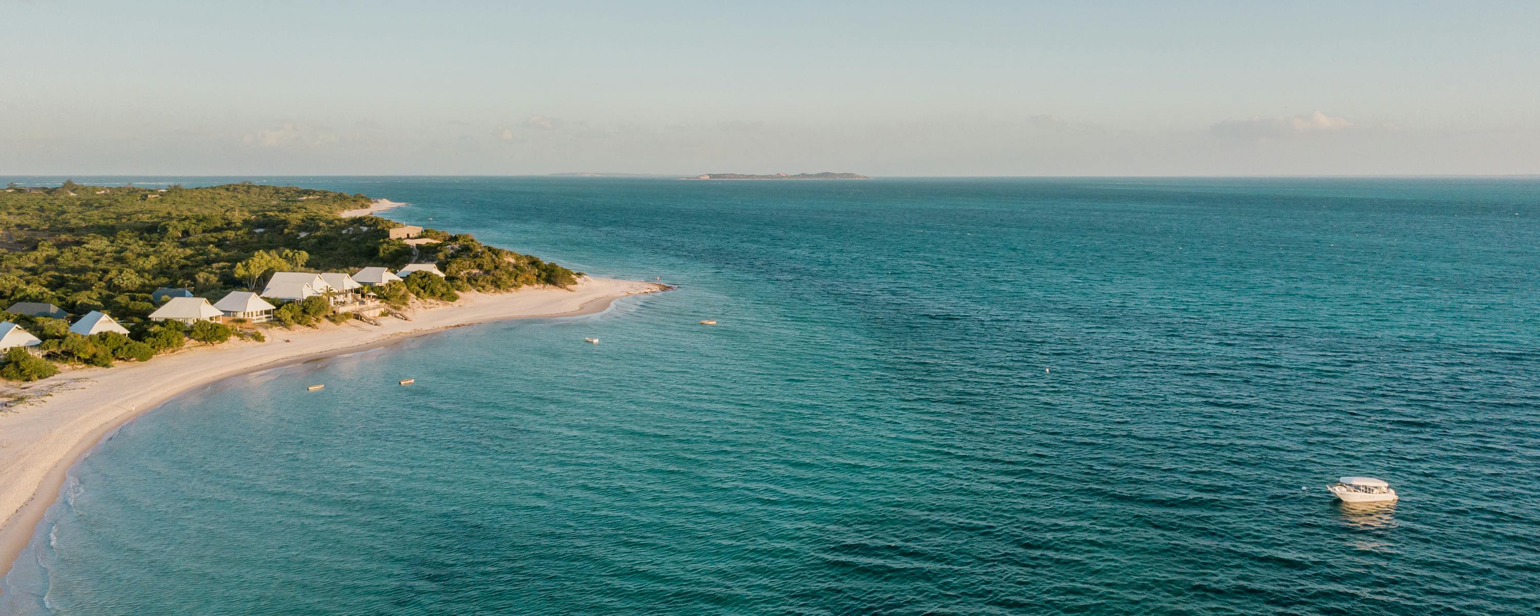Aerial view of a long sandy shoreline and turquoise sea, with low buildings and trees lining the coast below.