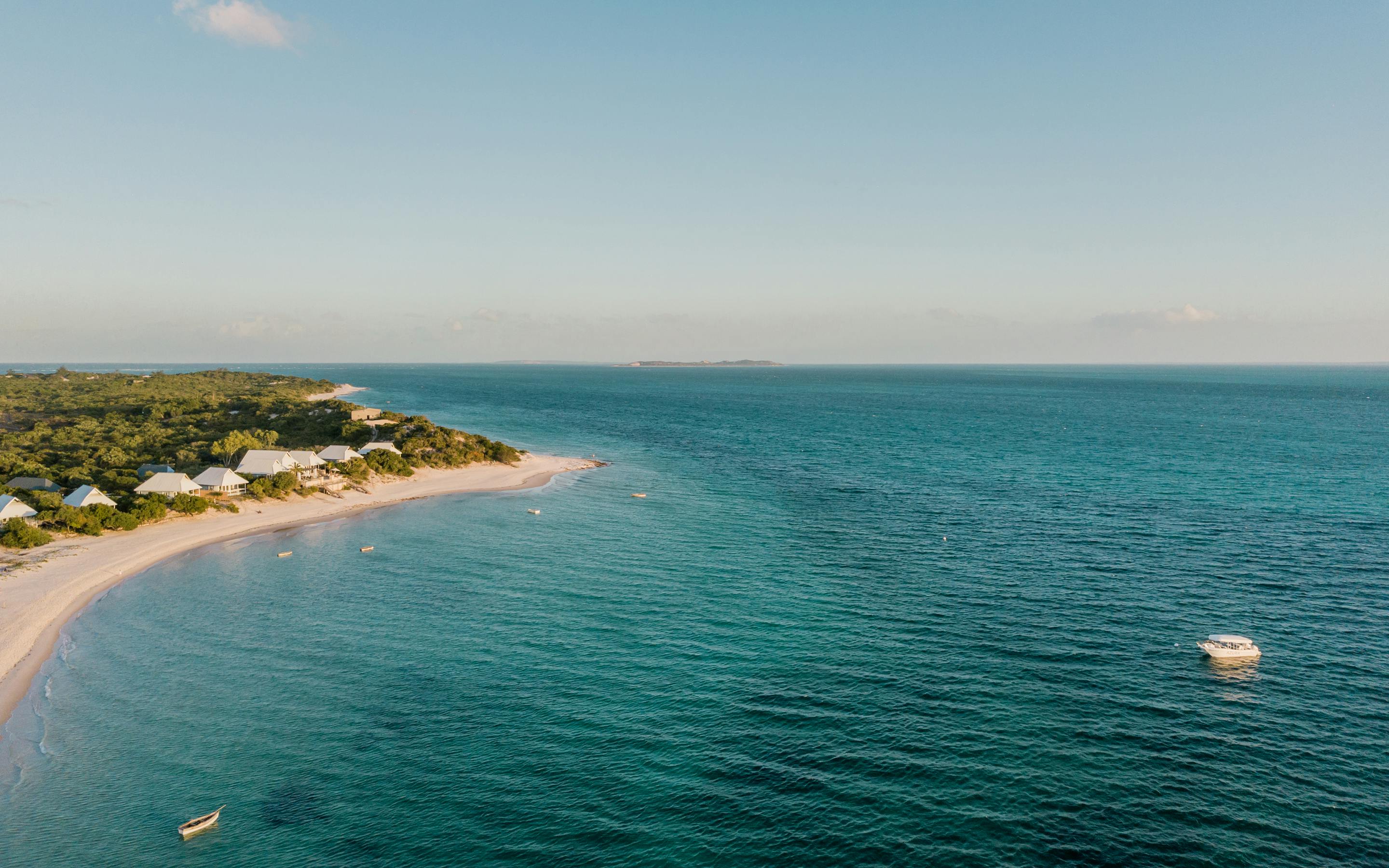 Aerial view of a long sandy shoreline and turquoise sea, with low buildings and trees lining the coast below.