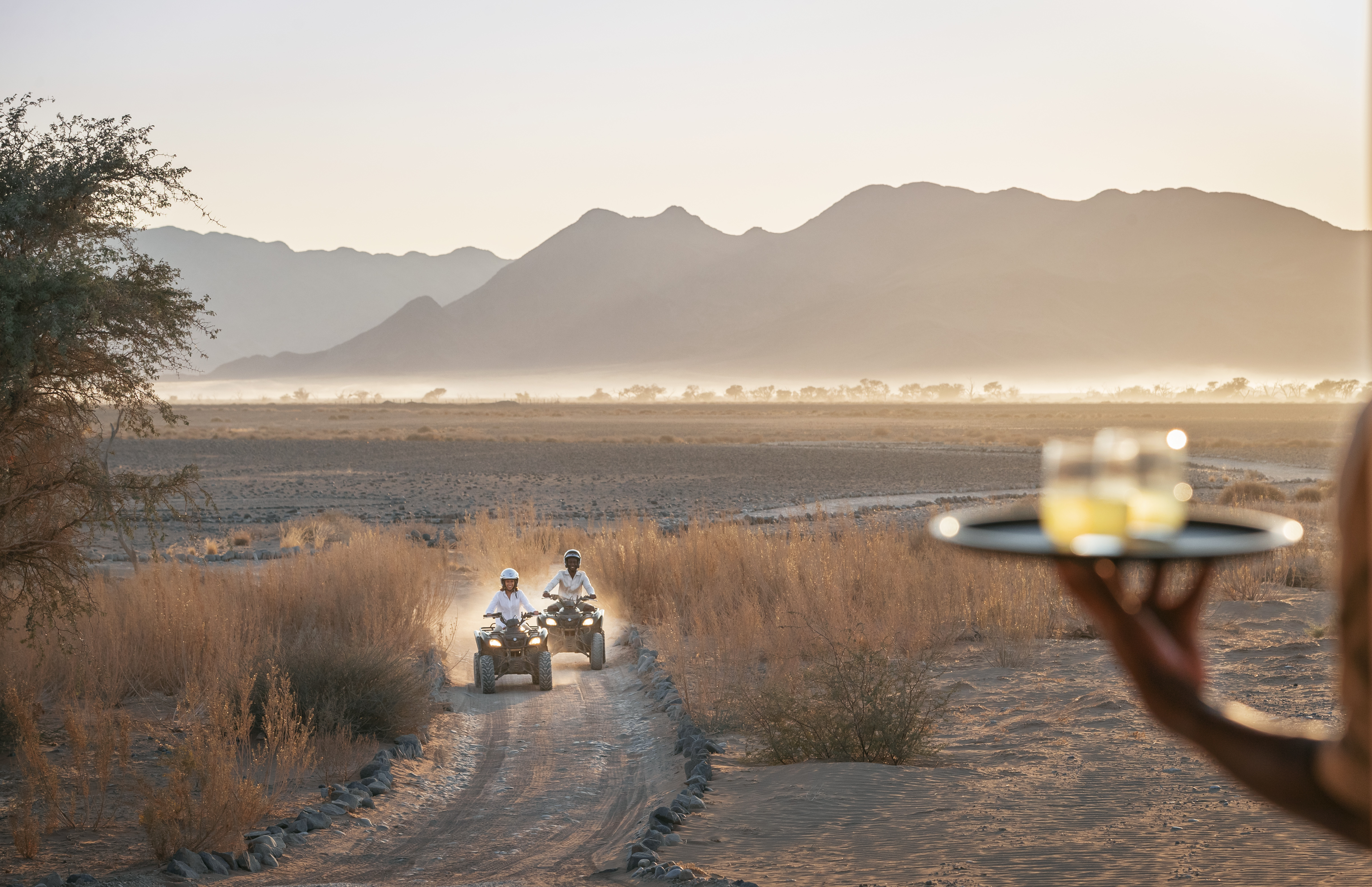 Two quad bikes drive along a sandy track at dusk while a tray of drinks is held up, with mountains in the distance.