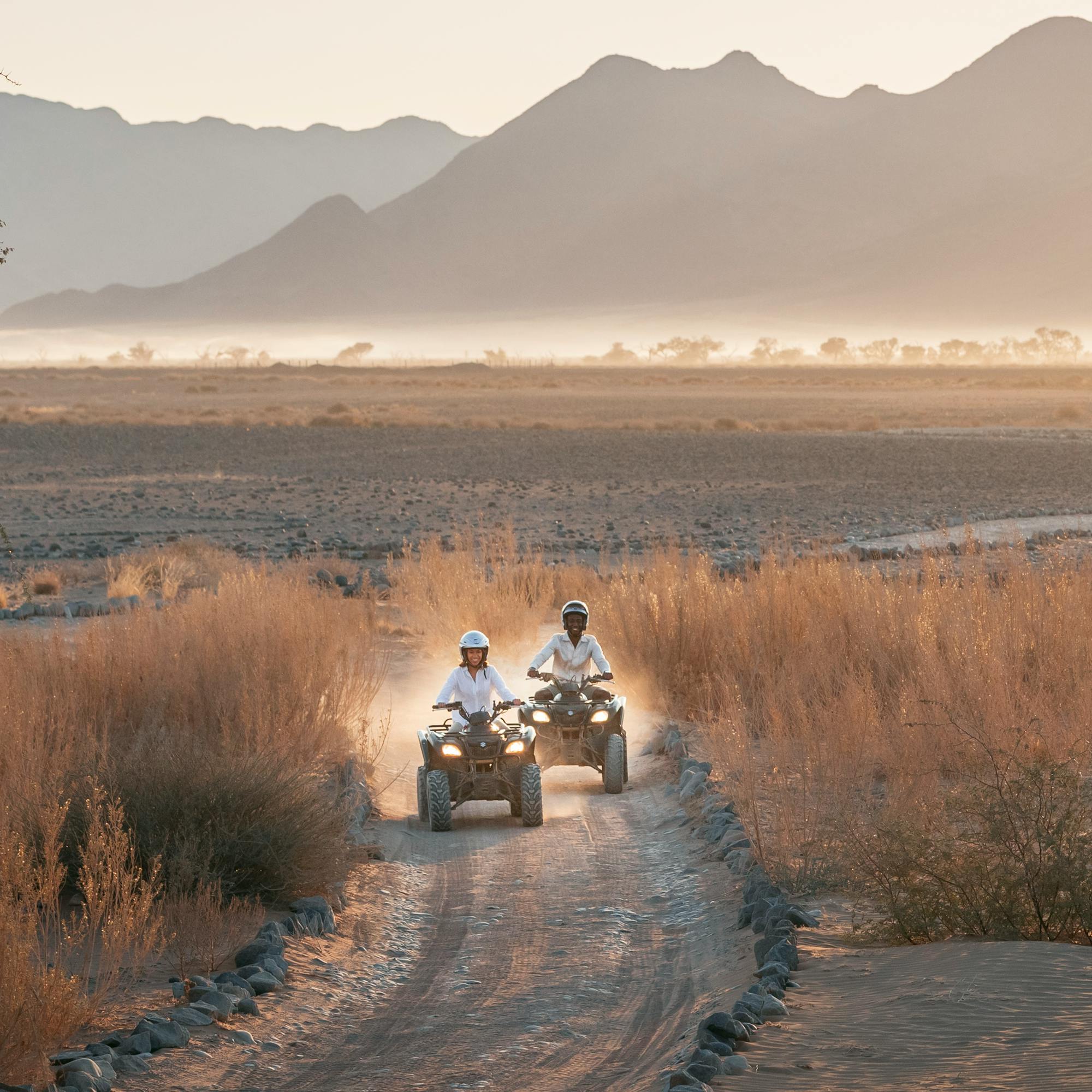 Two quad bikes drive along a sandy track at dusk while a tray of drinks is held up, with mountains in the distance.