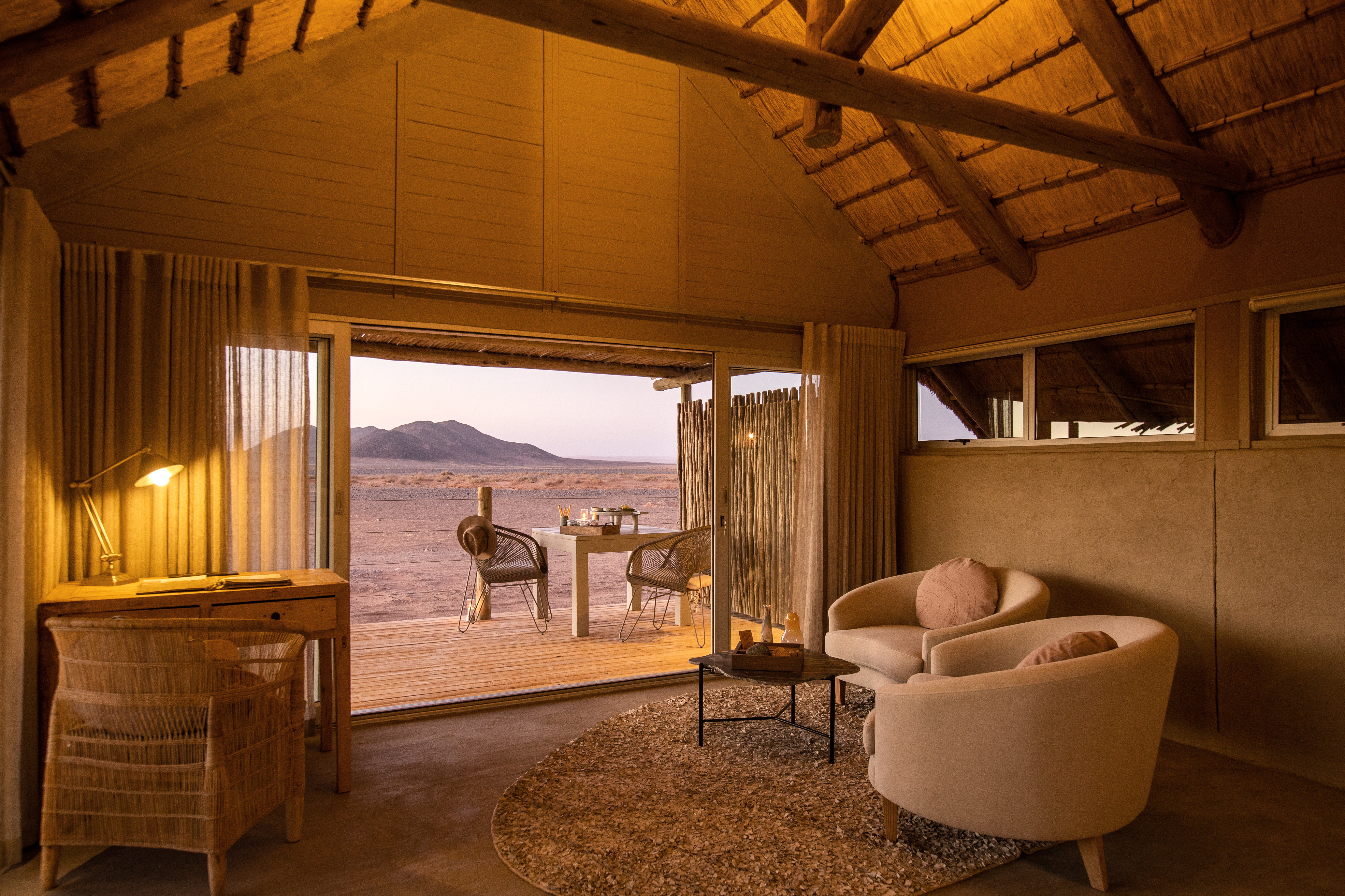 Lodge bedroom with draped canopy and seating opens to a shaded deck, framing distant dunes and mountains.