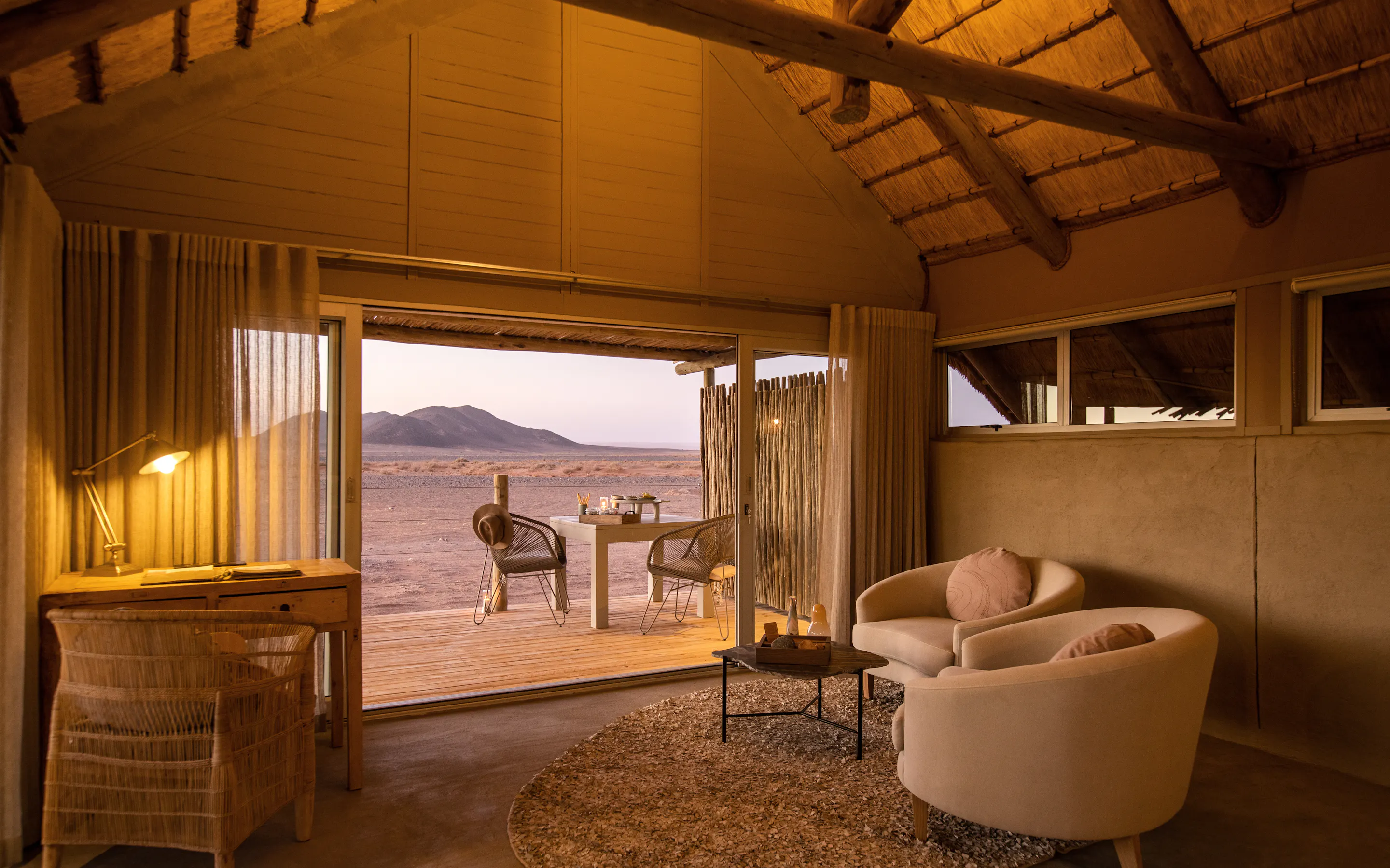 Lodge bedroom with draped canopy and seating opens to a shaded deck, framing distant dunes and mountains.