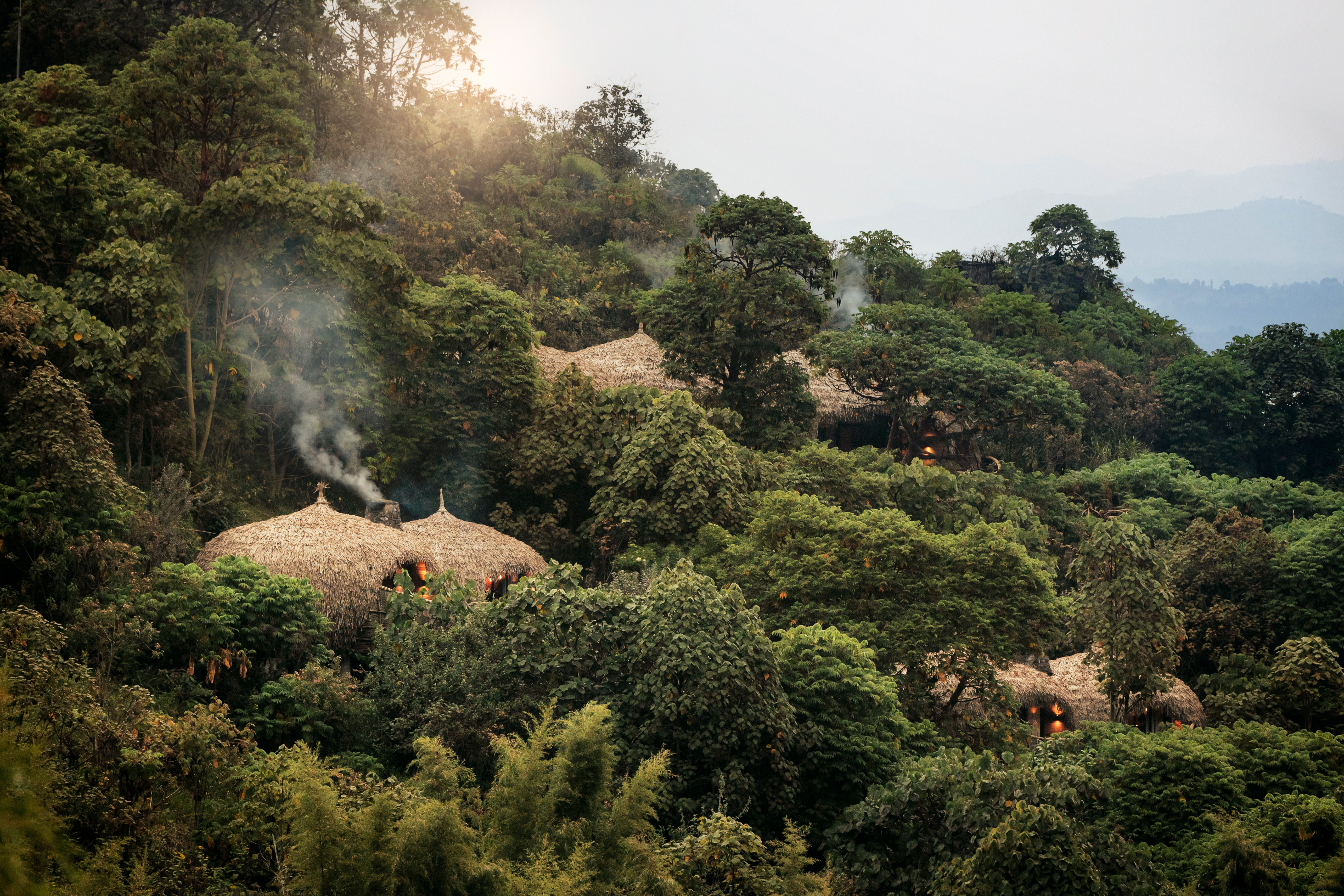 Thatched-roof huts sit among lush green hills, with warm morning light and mist rising above the treetops.