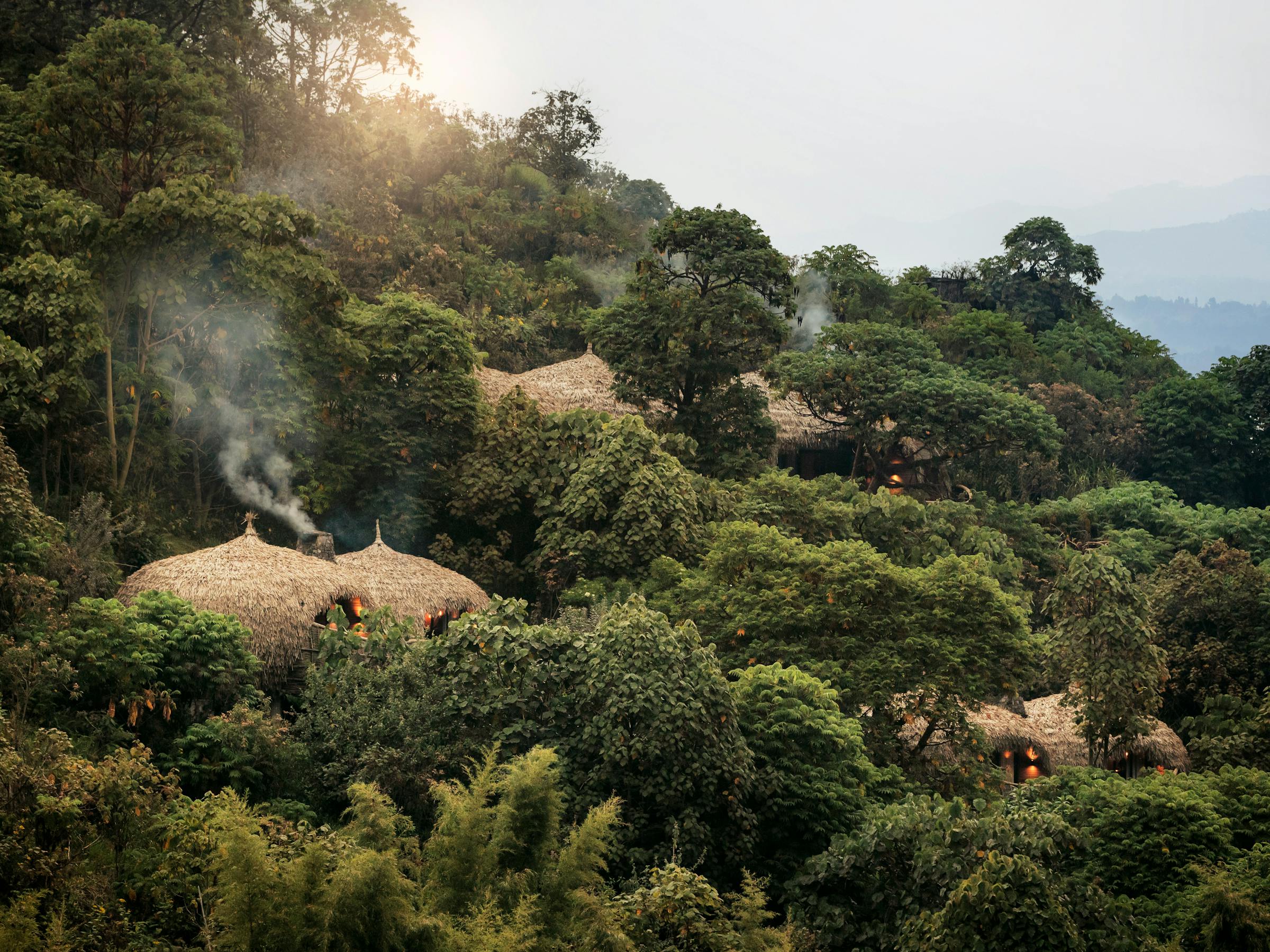 Thatched-roof huts sit among lush green hills, with warm morning light and mist rising above the treetops.