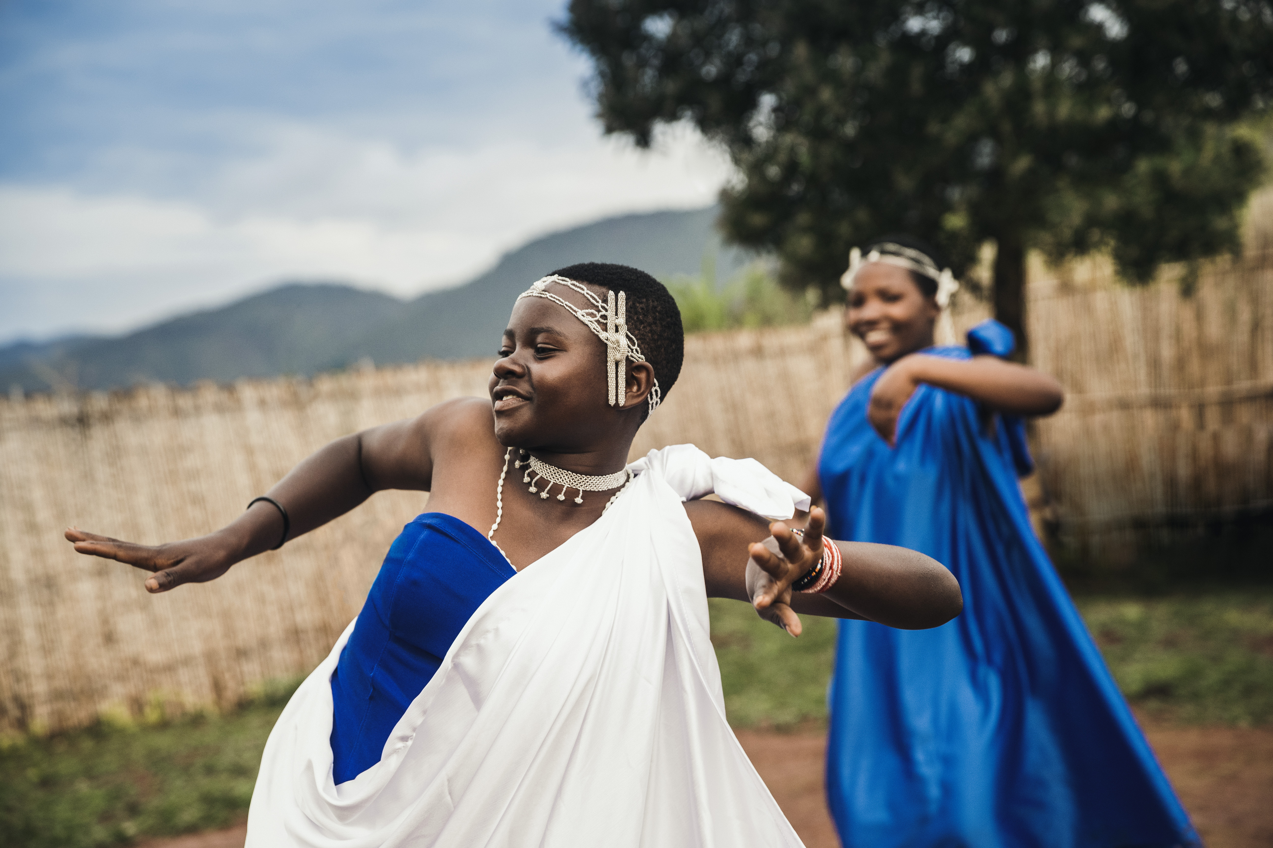 Two dancers in flowing white and blue traditional dresses perform outdoors, arms outstretched near a woven fence.
