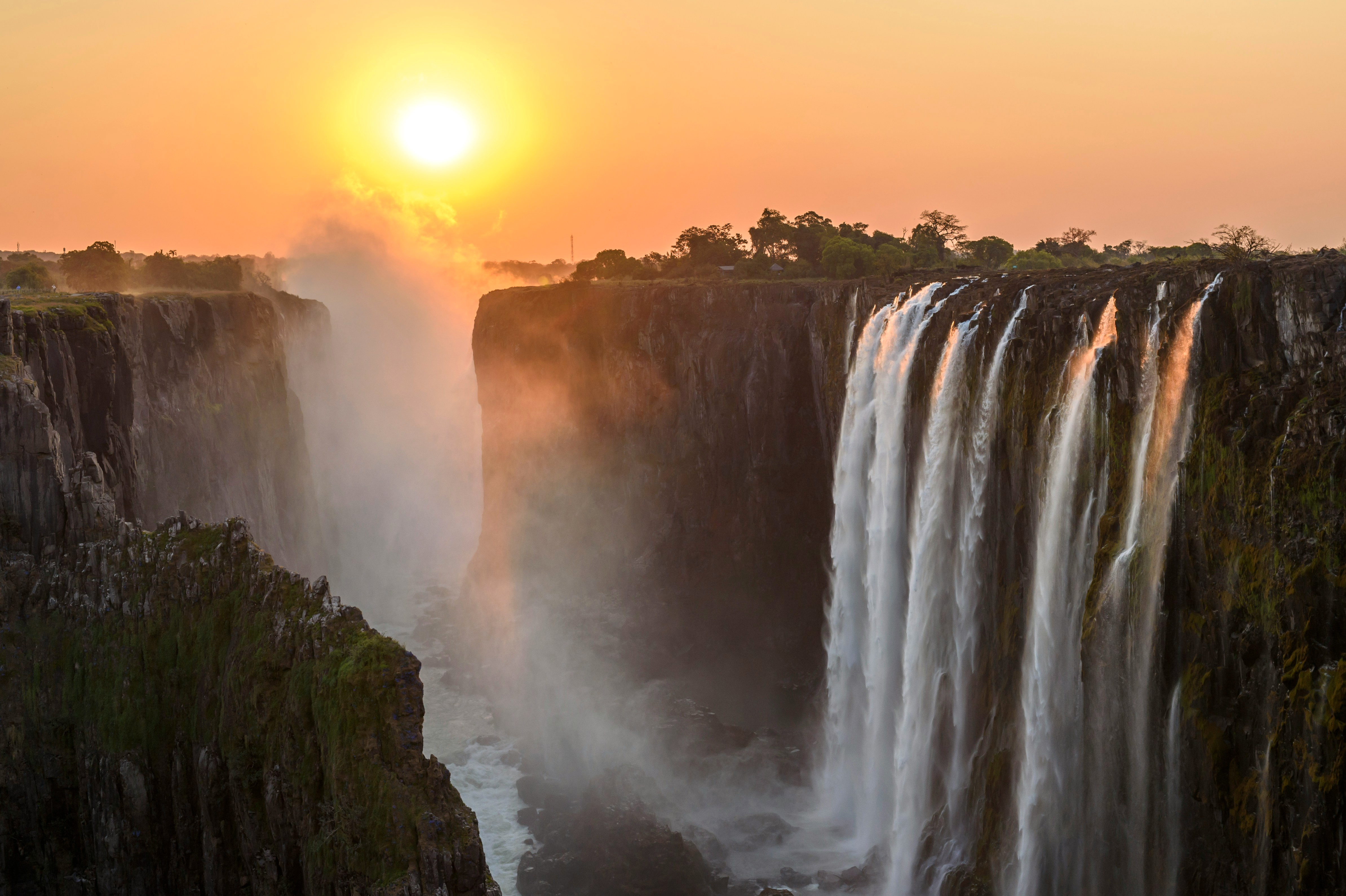 Sun sets over a broad waterfall as mist rises from the gorge, with orange sky, dark cliffs, and forest along the rim.