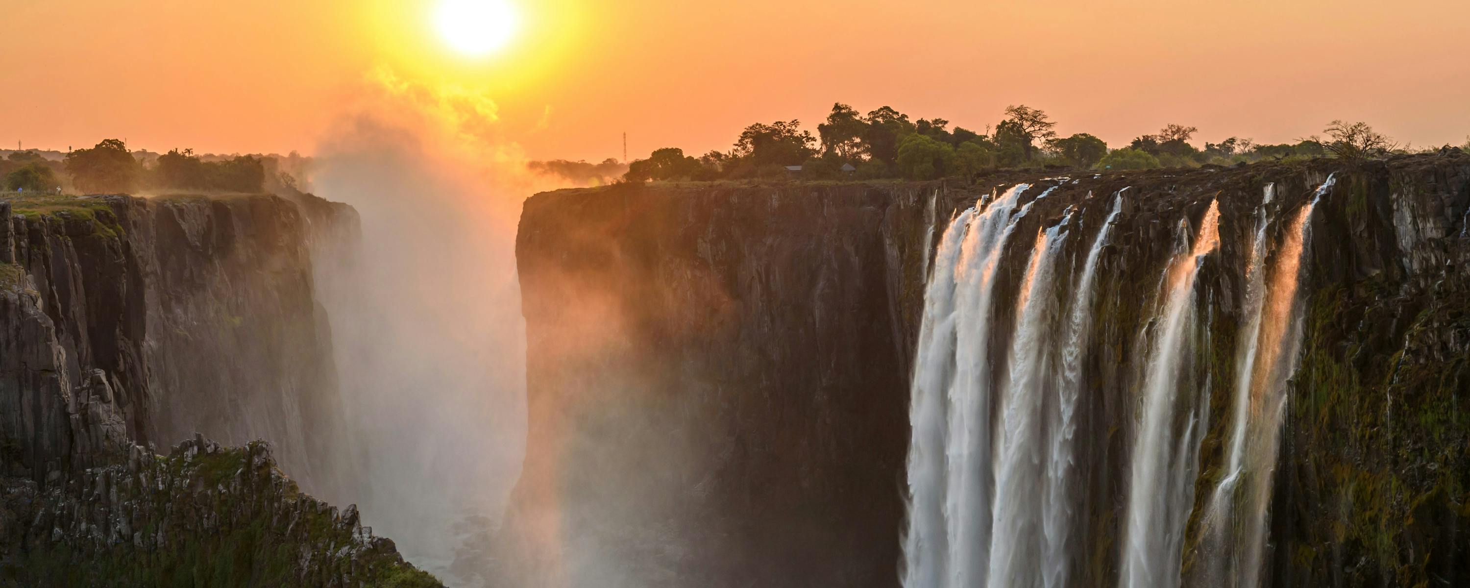 Sun sets over a broad waterfall as mist rises from the gorge, with orange sky, dark cliffs, and forest along the rim.