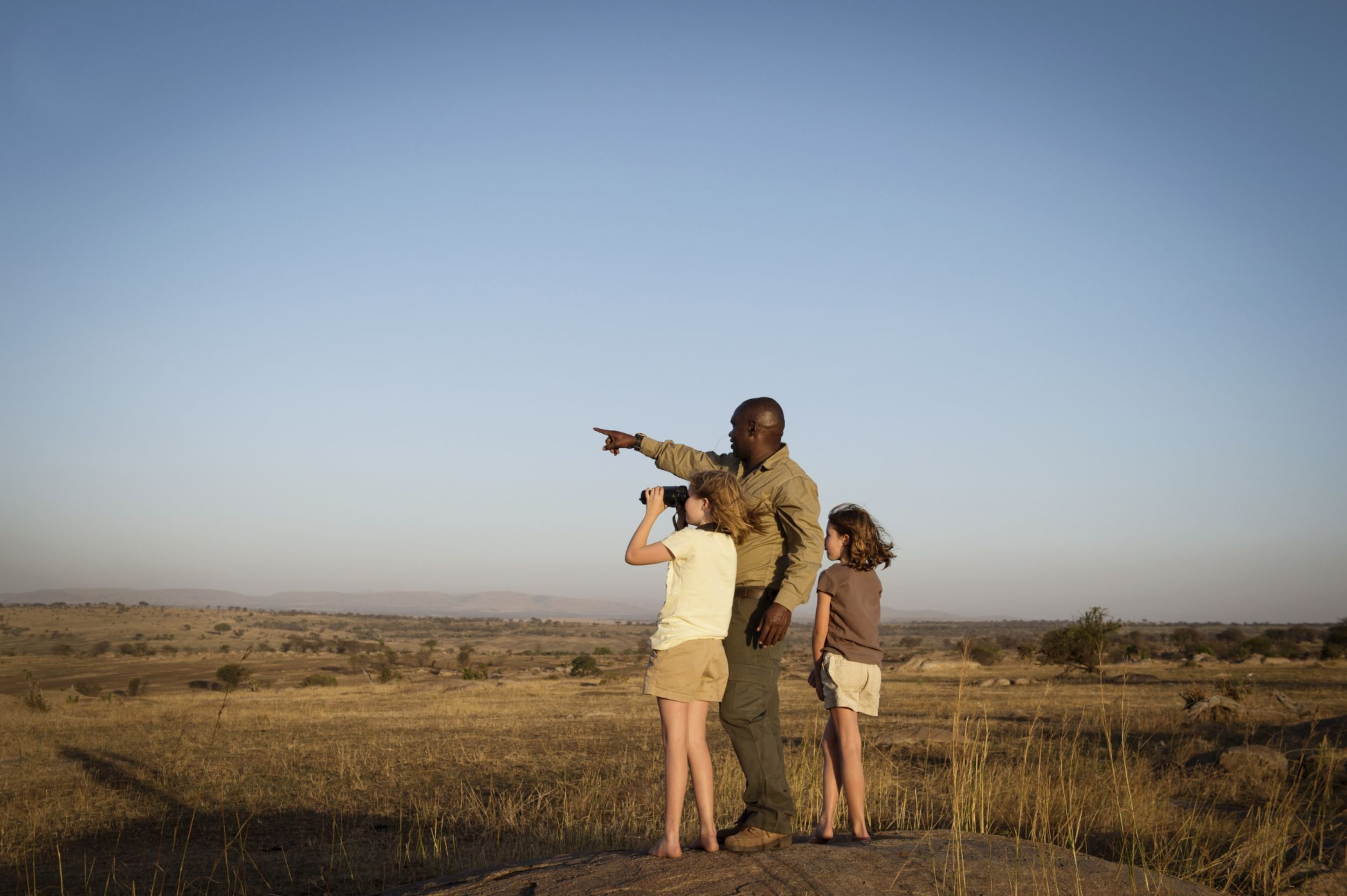Three people stand on a grassy ridge at sunset, one pointing while another looks through binoculars over the plain.