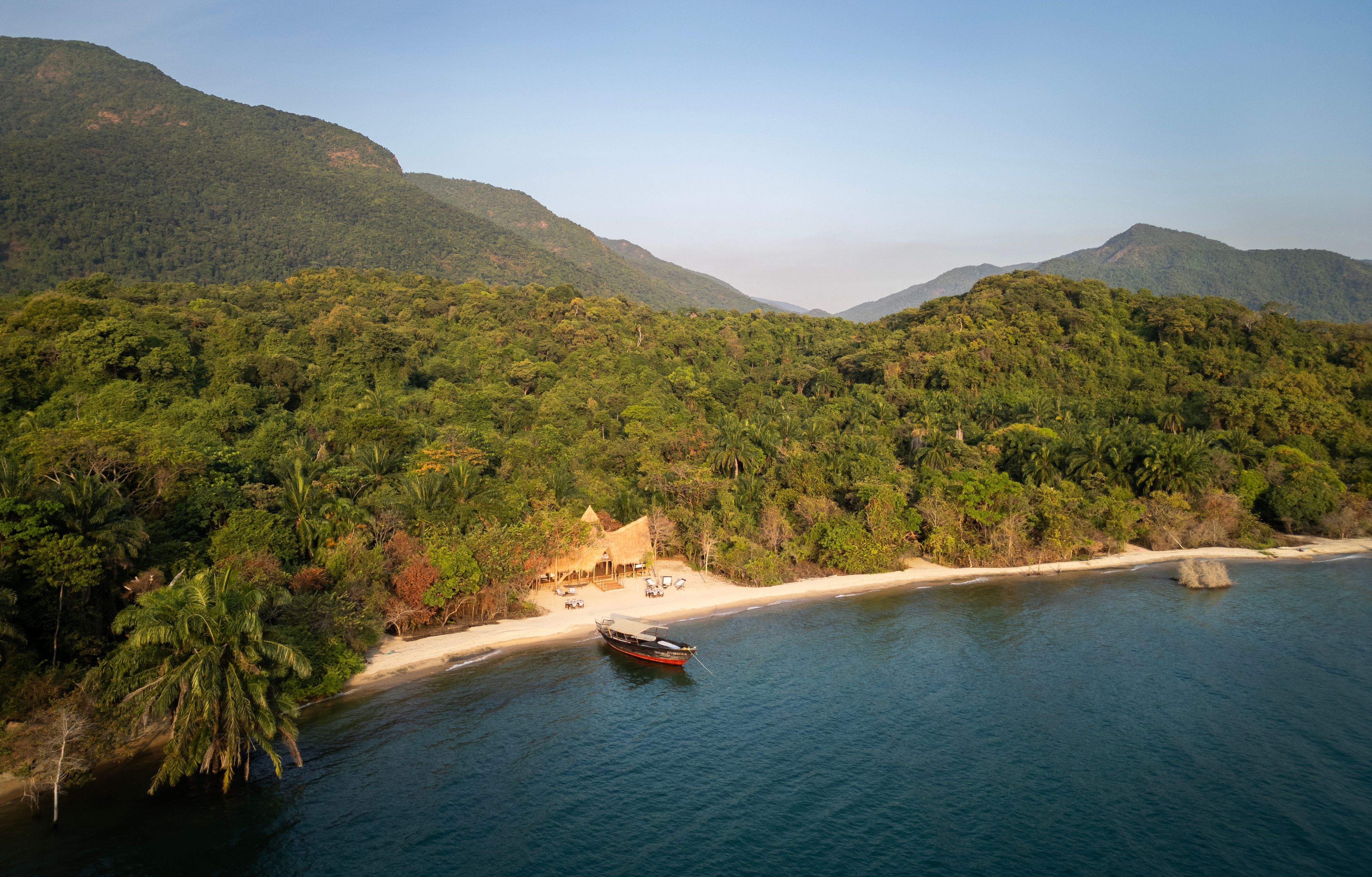 A high view shows a sandy beach and green forest meeting deep blue water, with a small boat near the quiet shore.