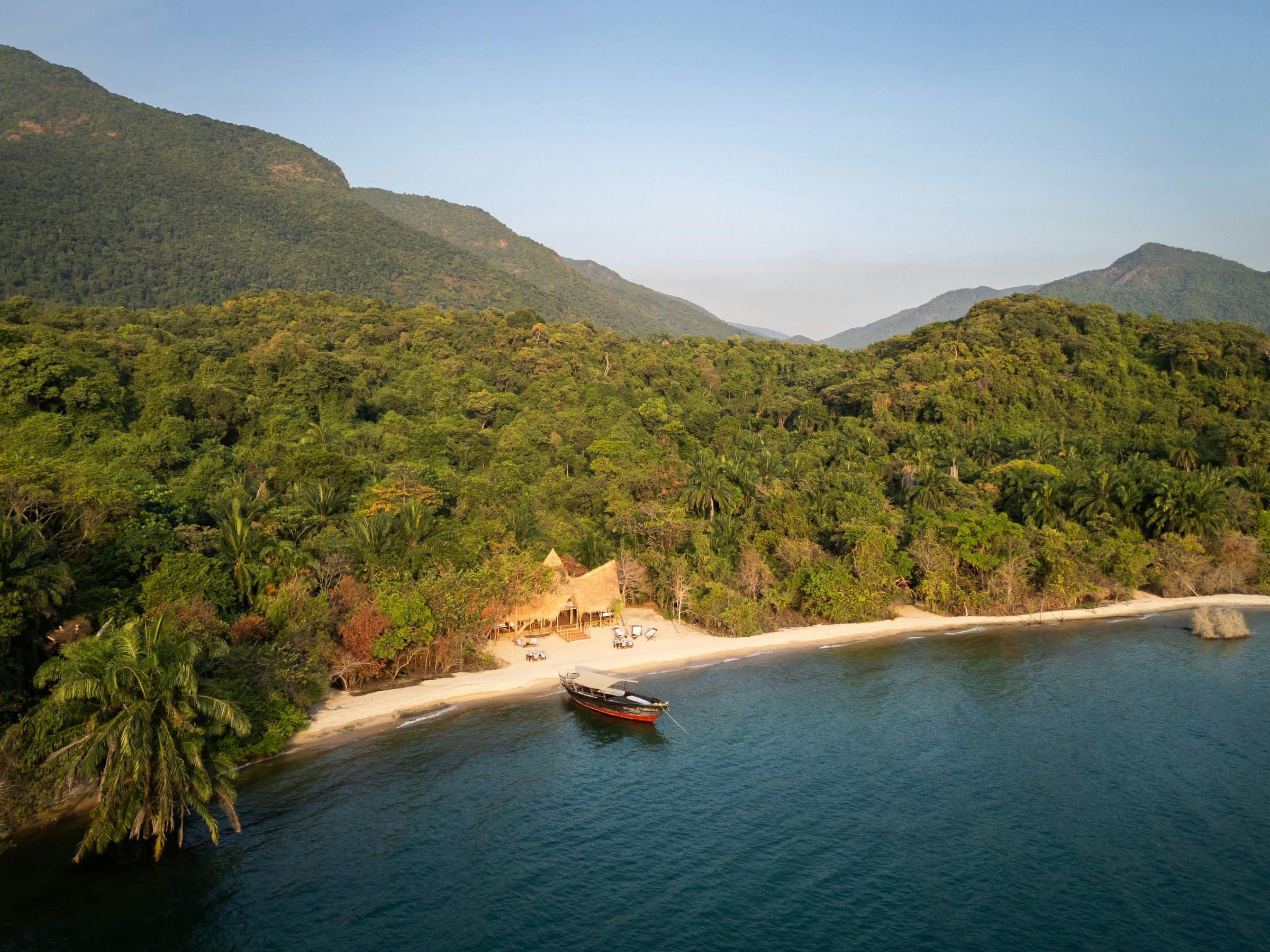 A high view shows a sandy beach and green forest meeting deep blue water, with a small boat near the quiet shore.