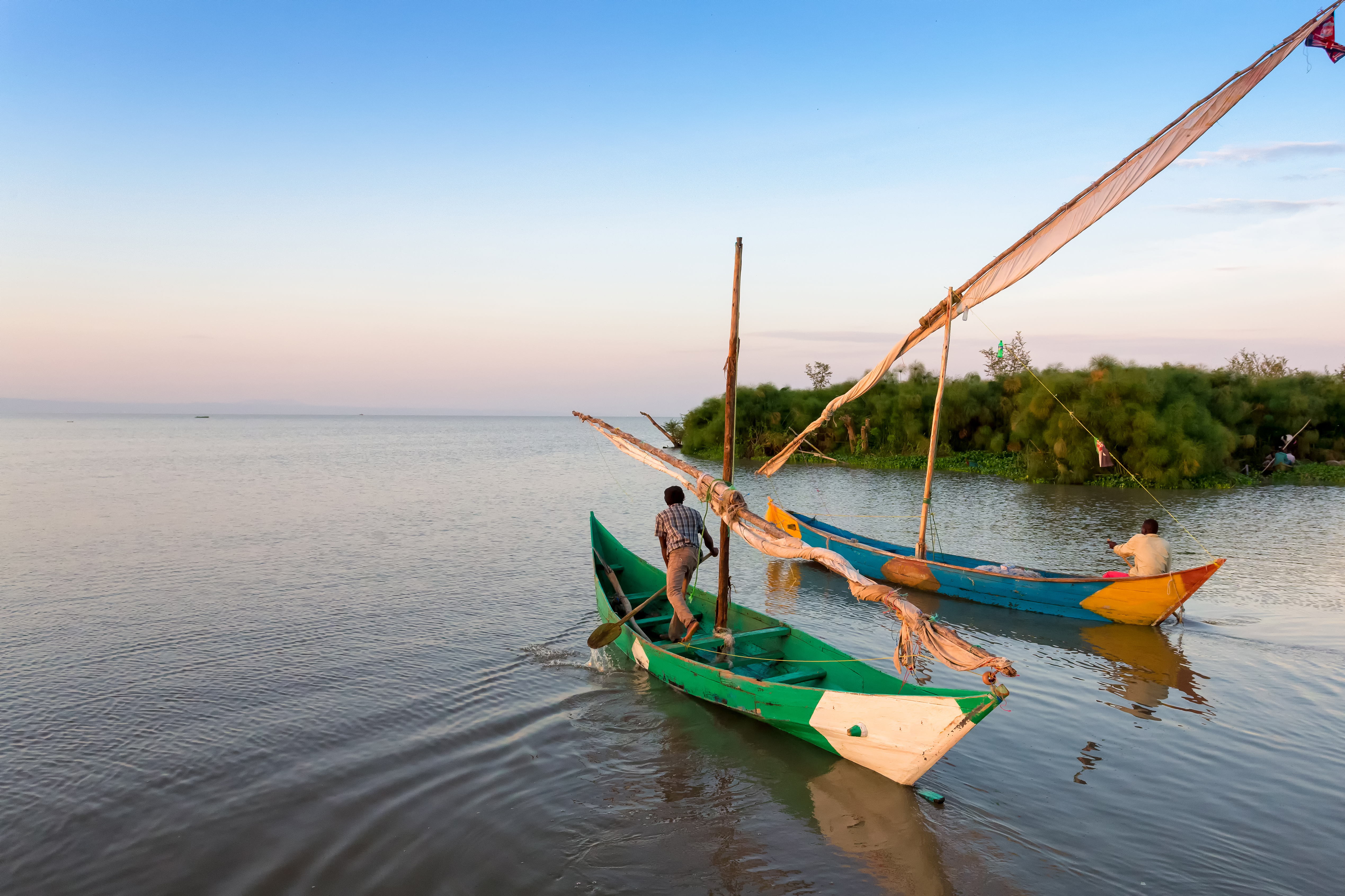 A wooden sailboat rests on calm water at sunset, with two people on deck and a green shoreline in the distance.