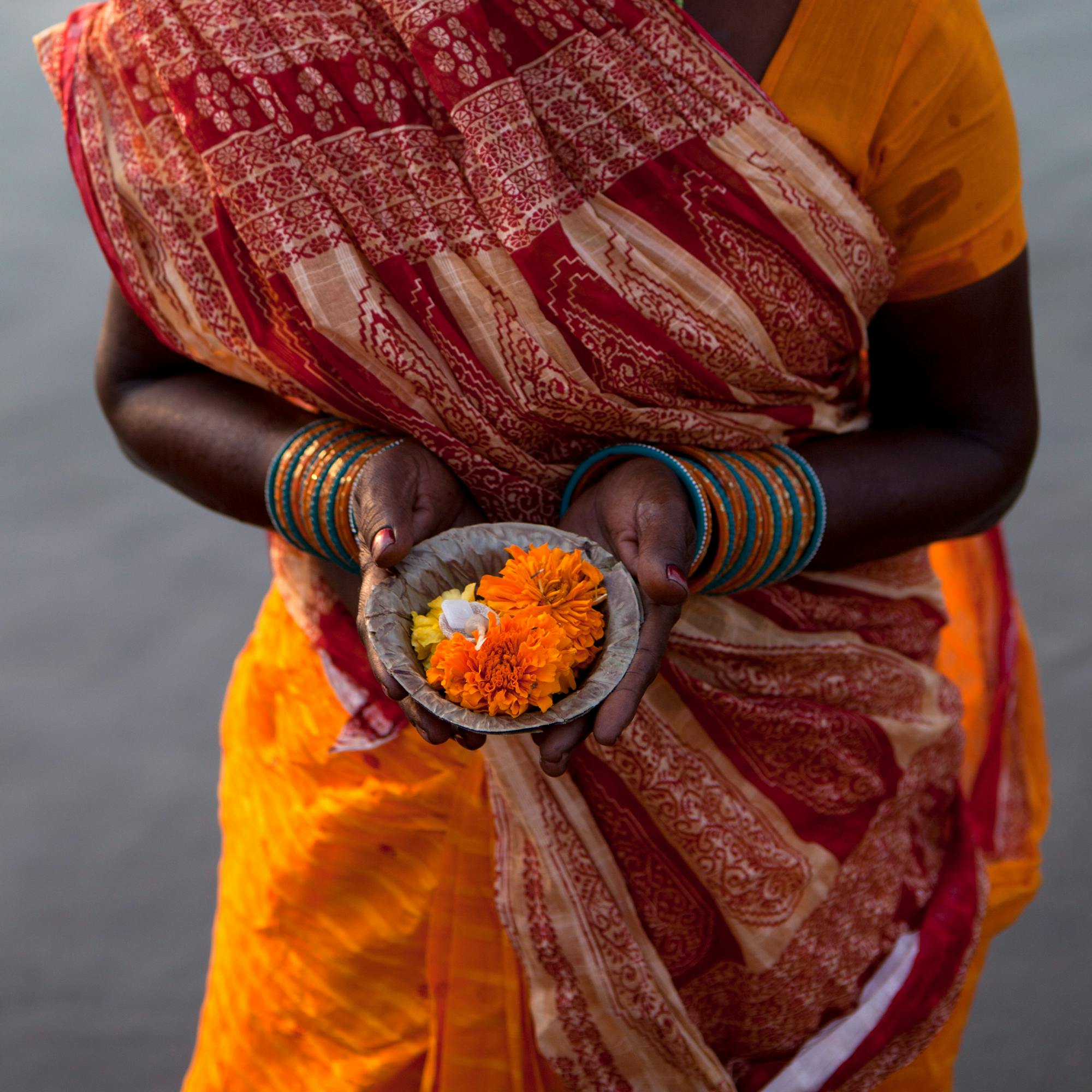 Person in a patterned sari holds a small bowl filled with marigold flowers.