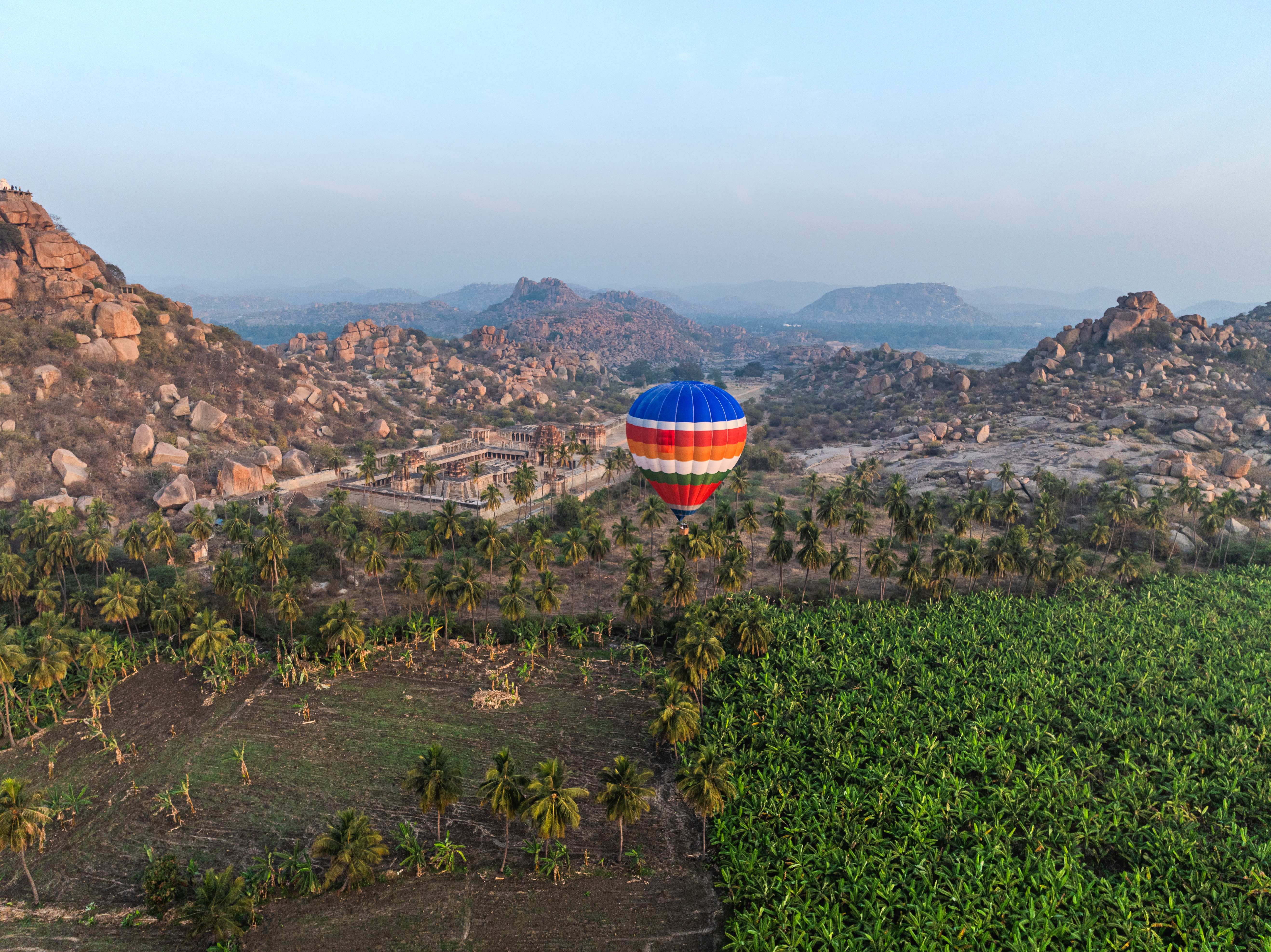 Hot air balloon floats above rocky hills and green fields under a hazy sky.