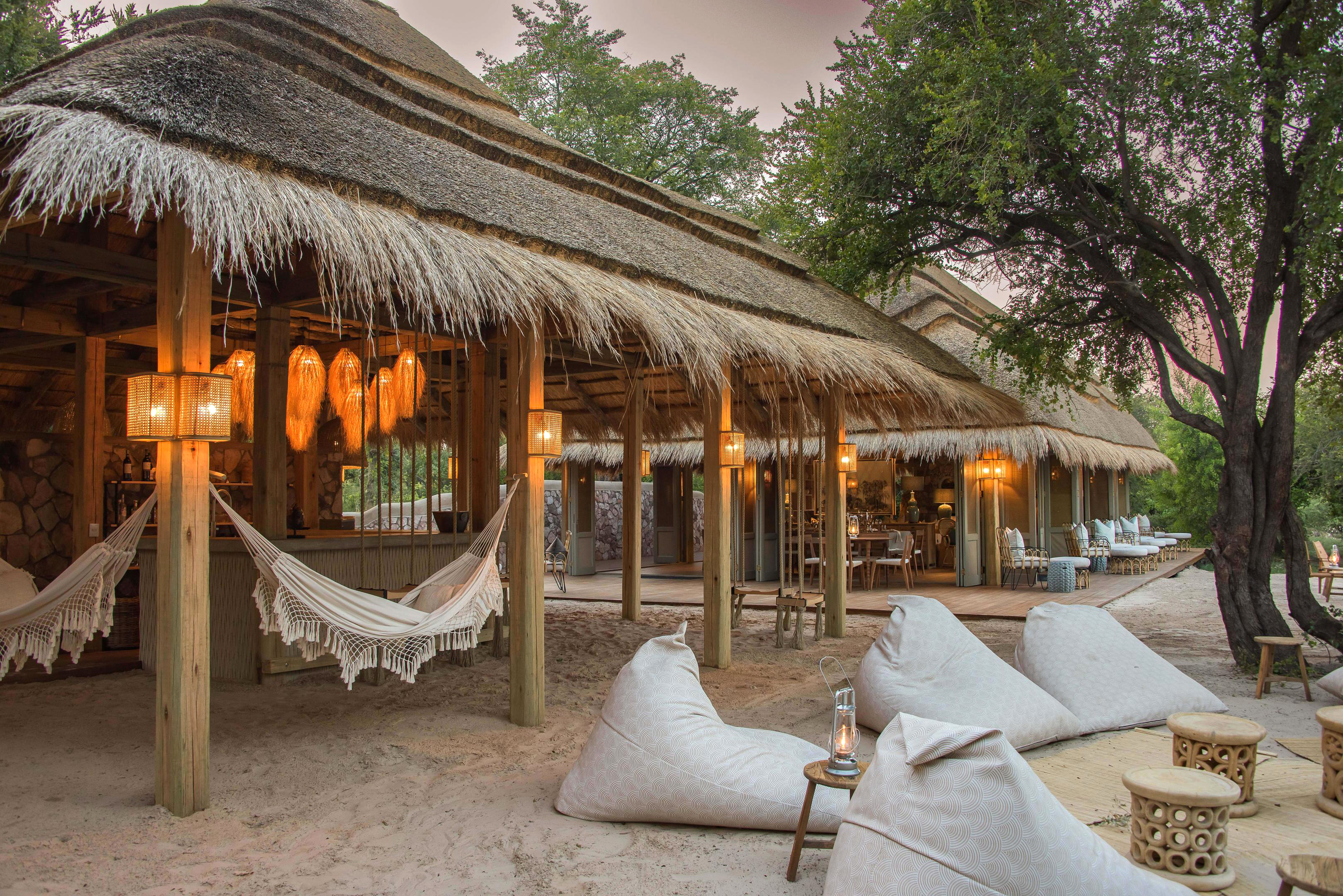 Thatched lodge veranda with hammocks, lanterns, and beanbag seats overlooks sandy ground under shade trees.