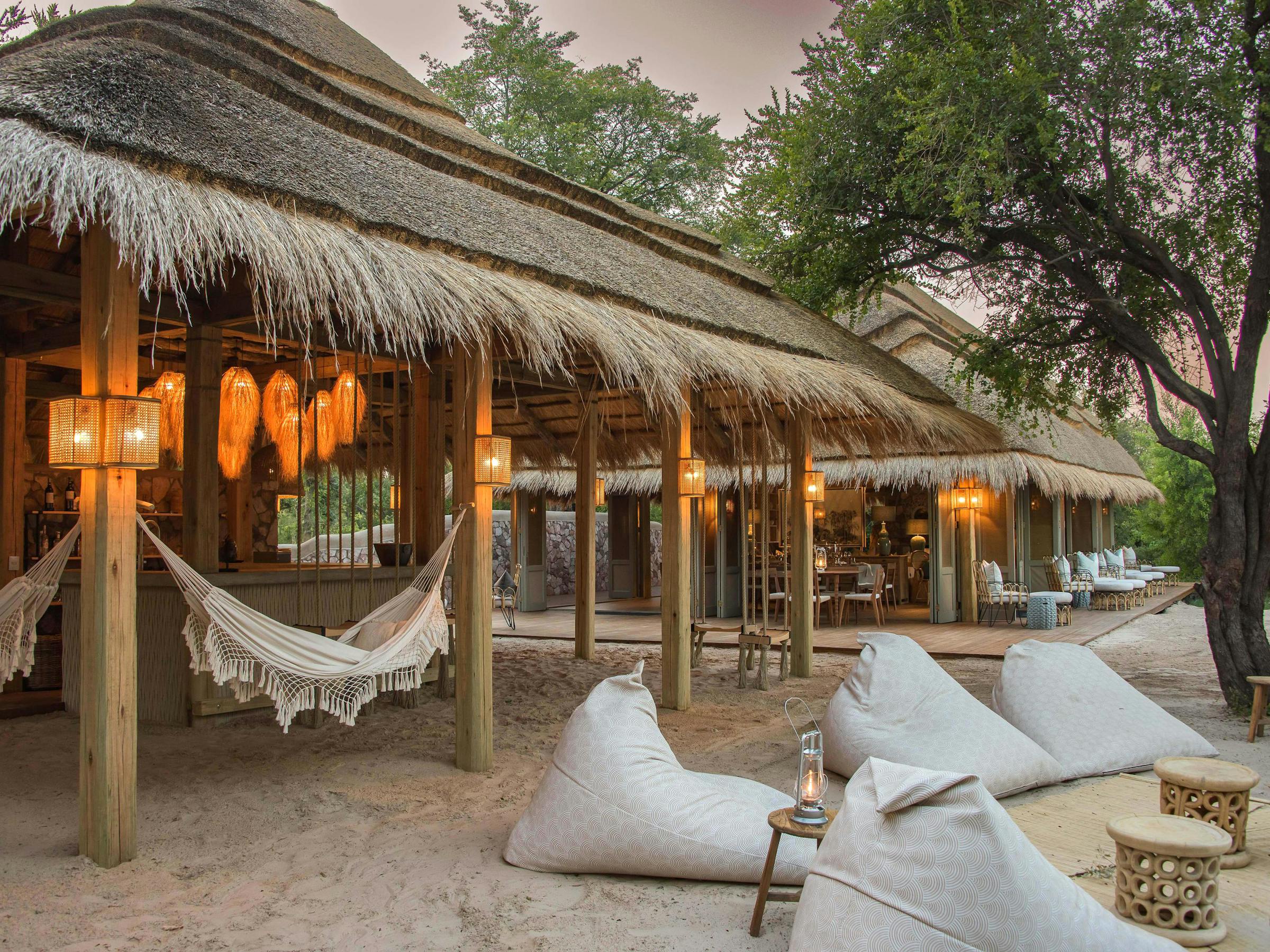 Thatched lodge veranda with hammocks, lanterns, and beanbag seats overlooks sandy ground under shade trees.