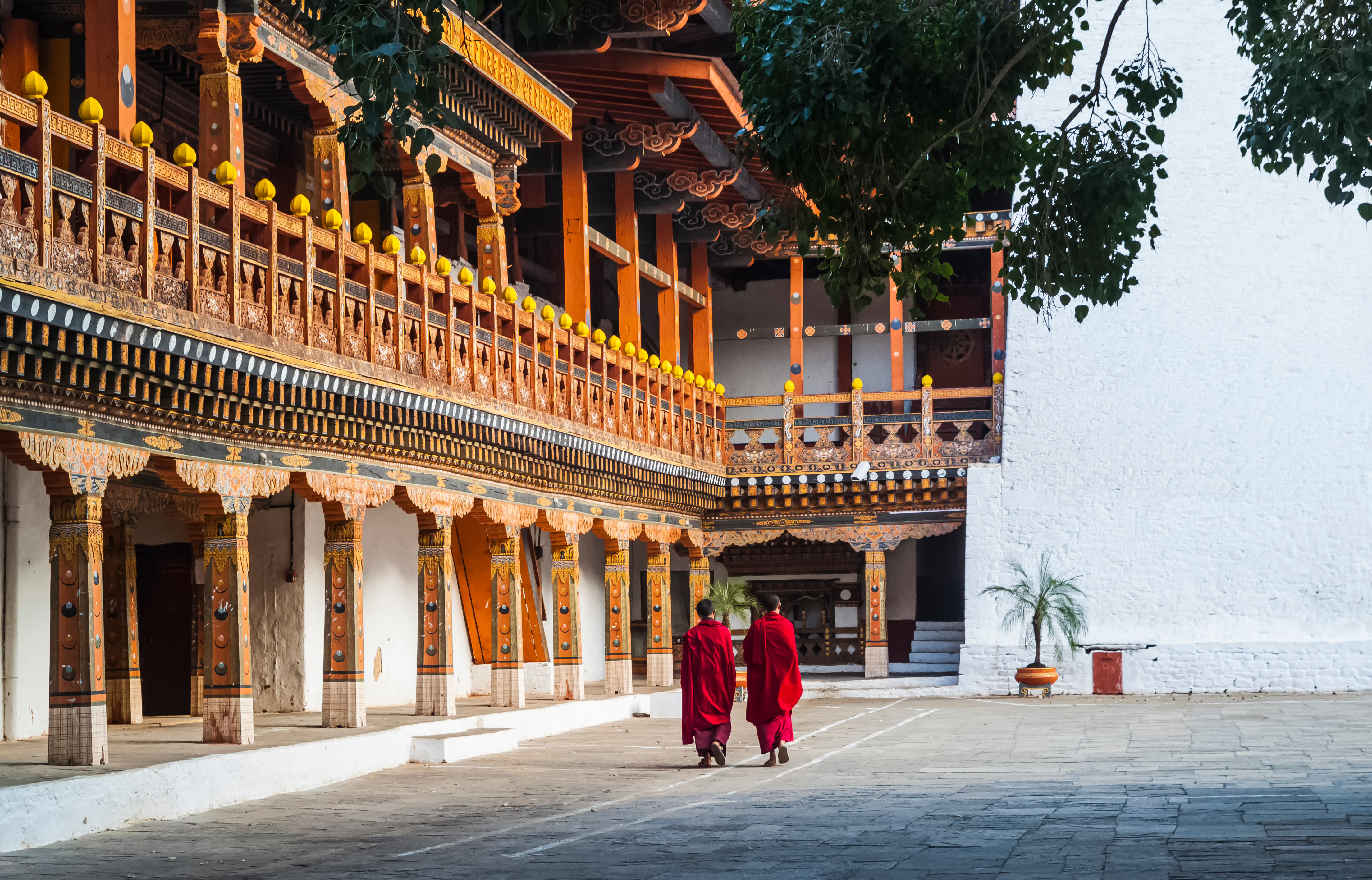 Two people in red robes walk across a monastery courtyard beside ornate wooden balconies.