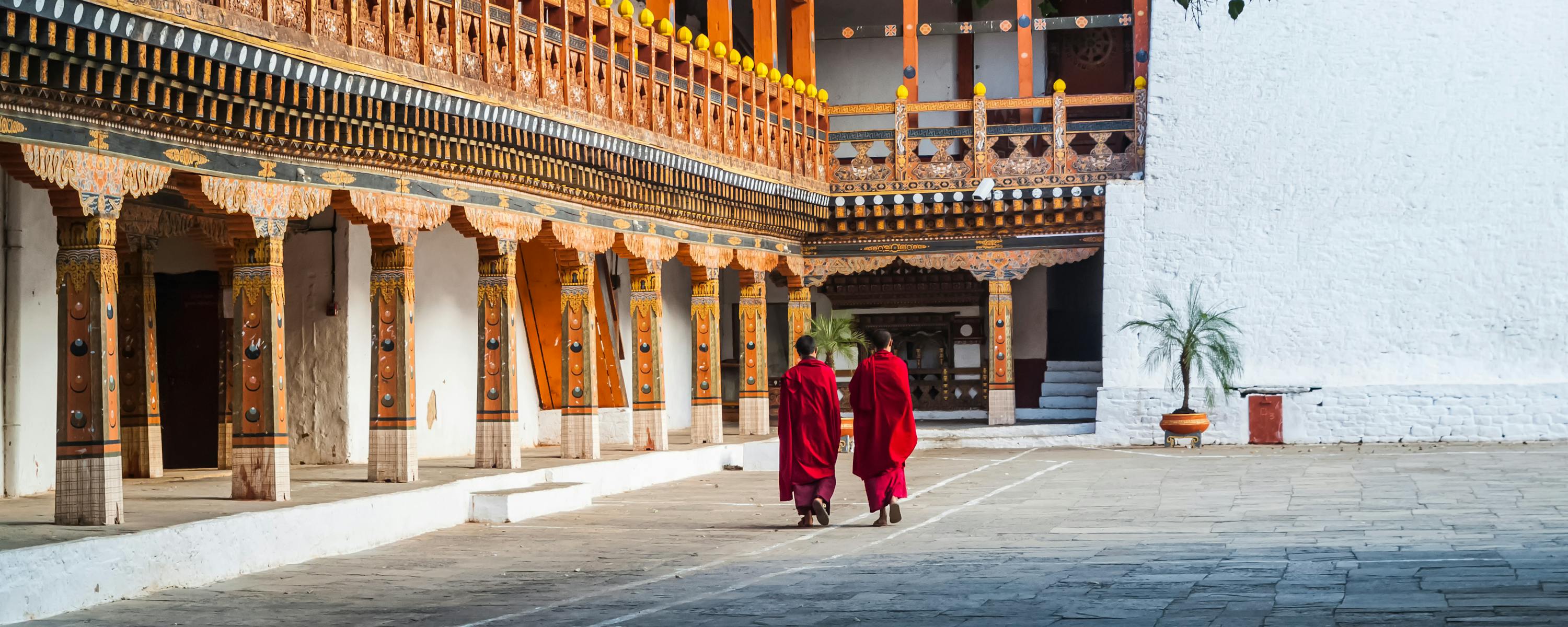 Two people in red robes walk across a monastery courtyard beside ornate wooden balconies.