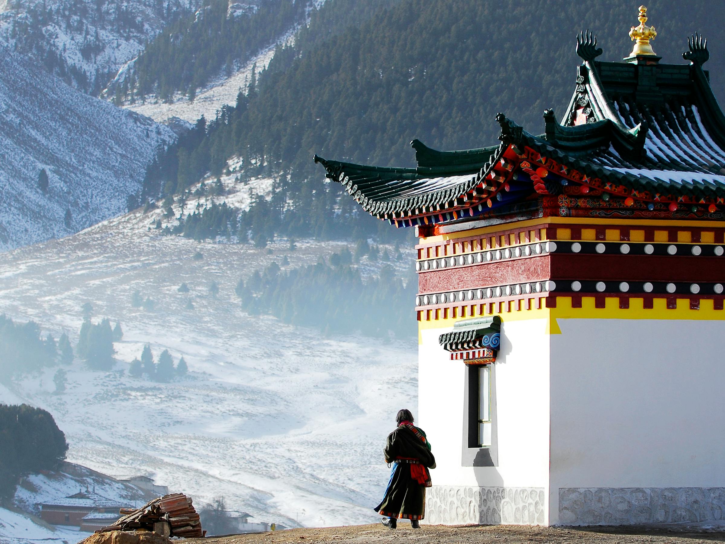 Person walks past a temple building with snowy mountains and a valley beyond.
