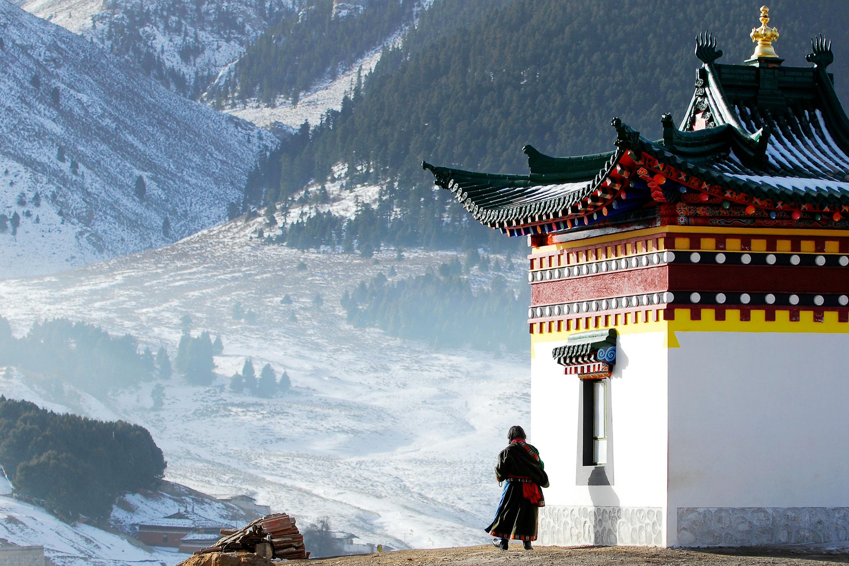 Person walks past a temple building with snowy mountains and a valley beyond.
