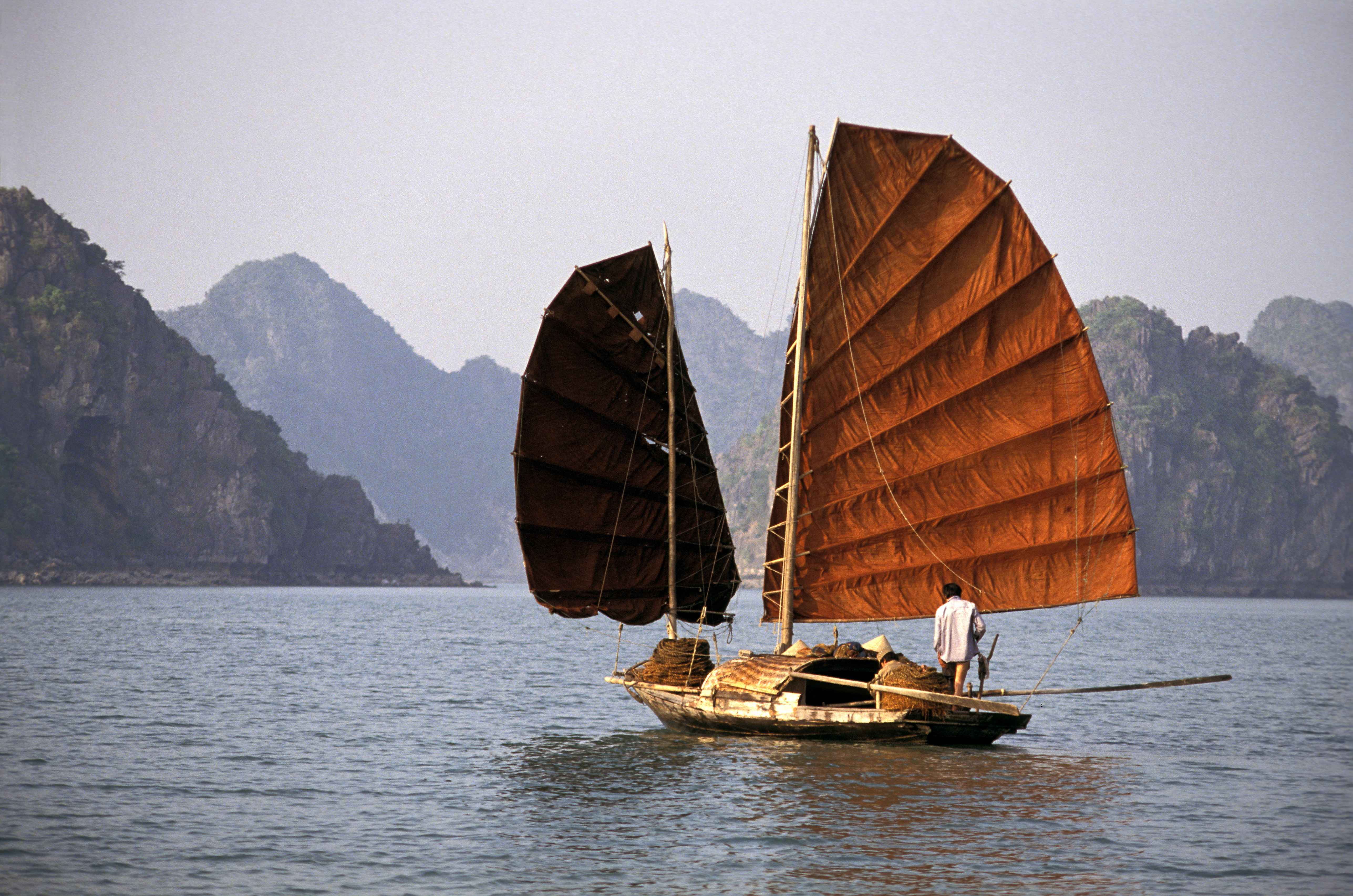 Traditional sailboat with red sails glides on calm water with limestone cliffs in the background.