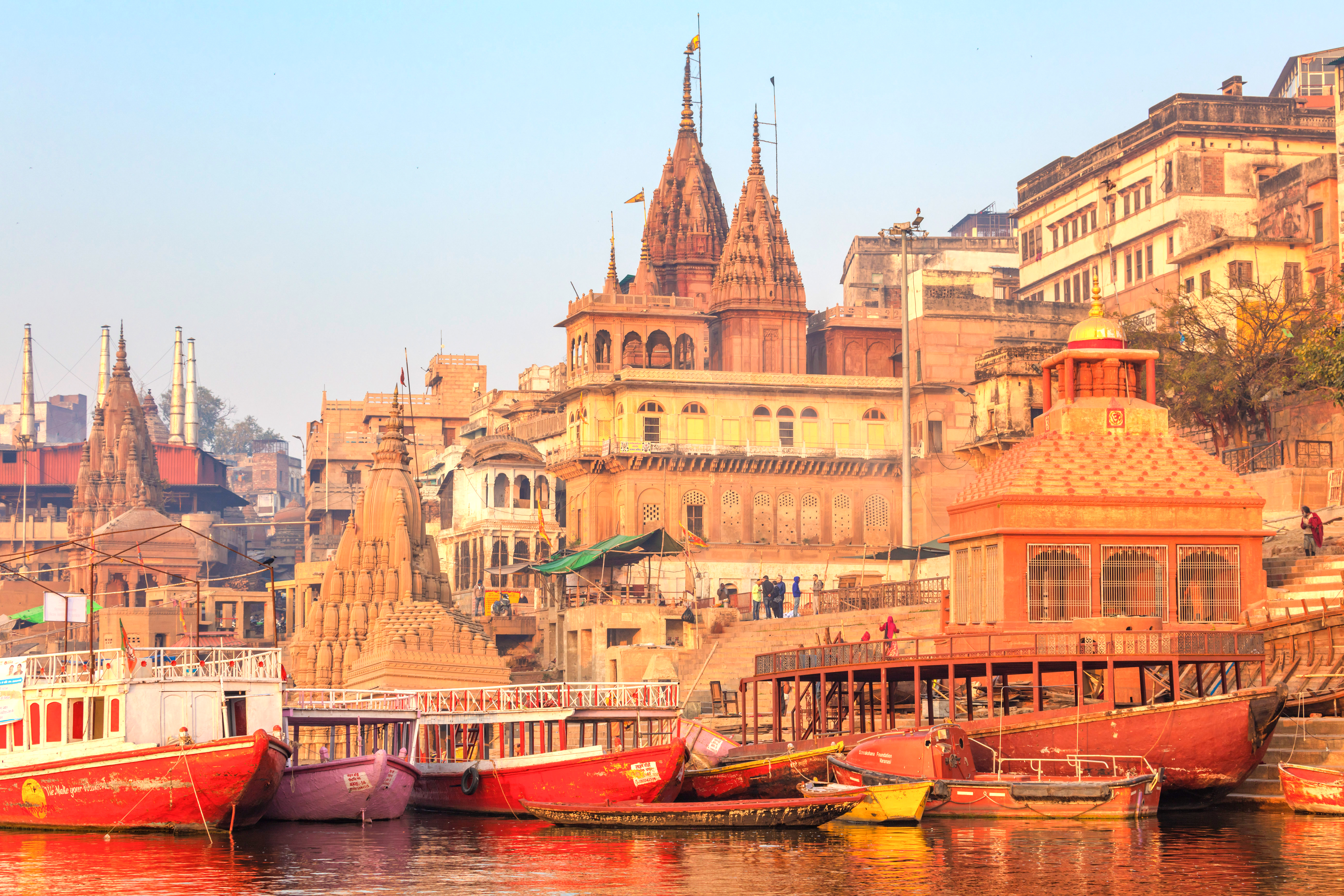 Boats line a riverbank crowded with historic buildings and temple spires at sunrise.