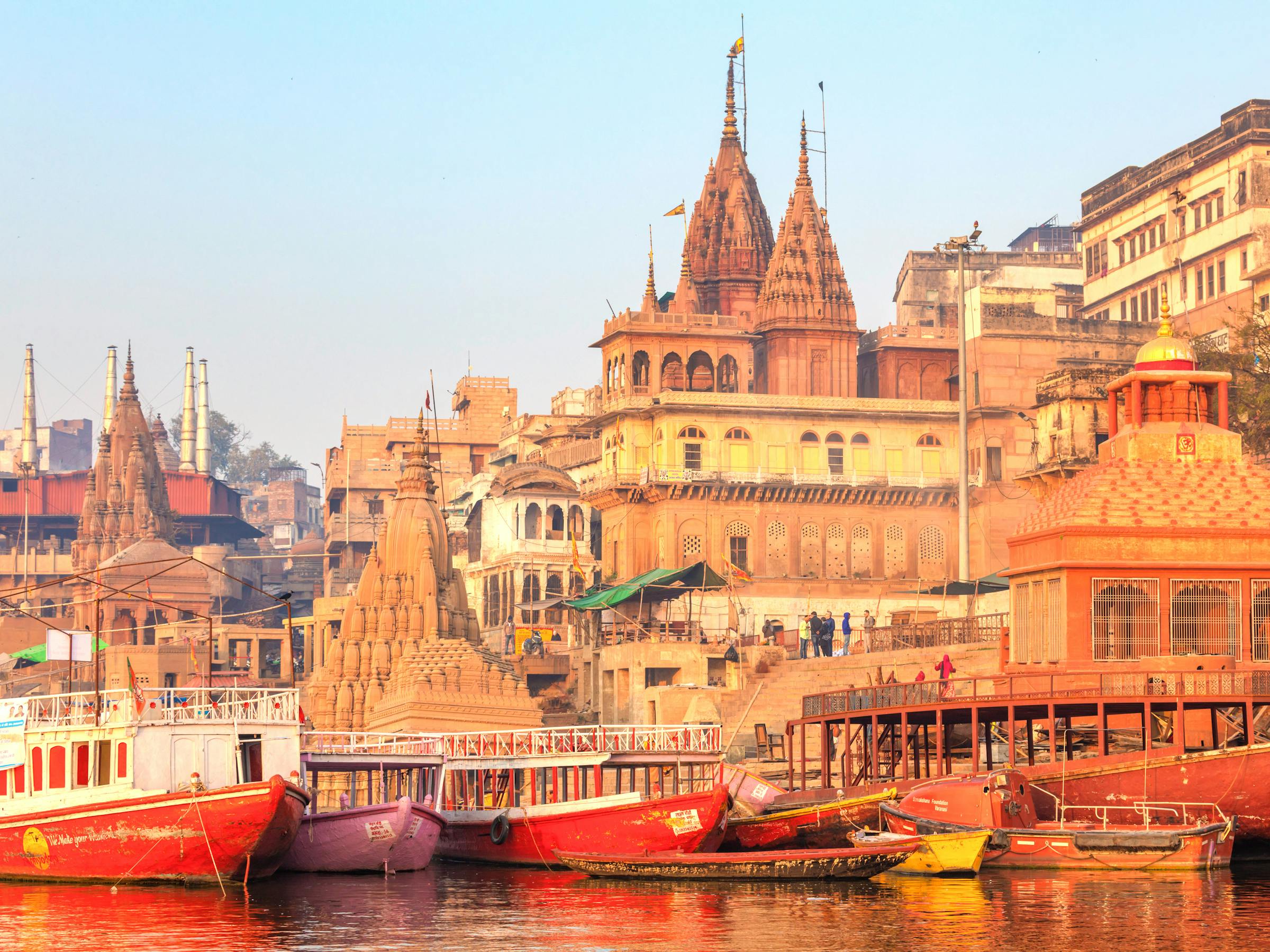 Boats line a riverbank crowded with historic buildings and temple spires at sunrise.