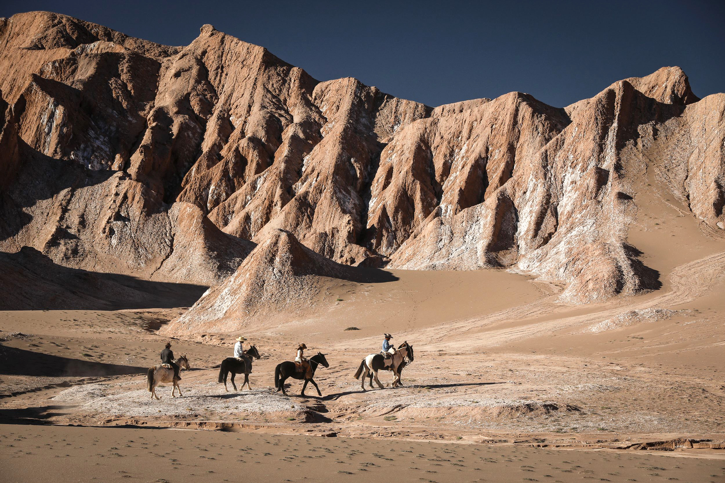 Horseback riders cross a sandy valley beneath tall rocky cliffs.
