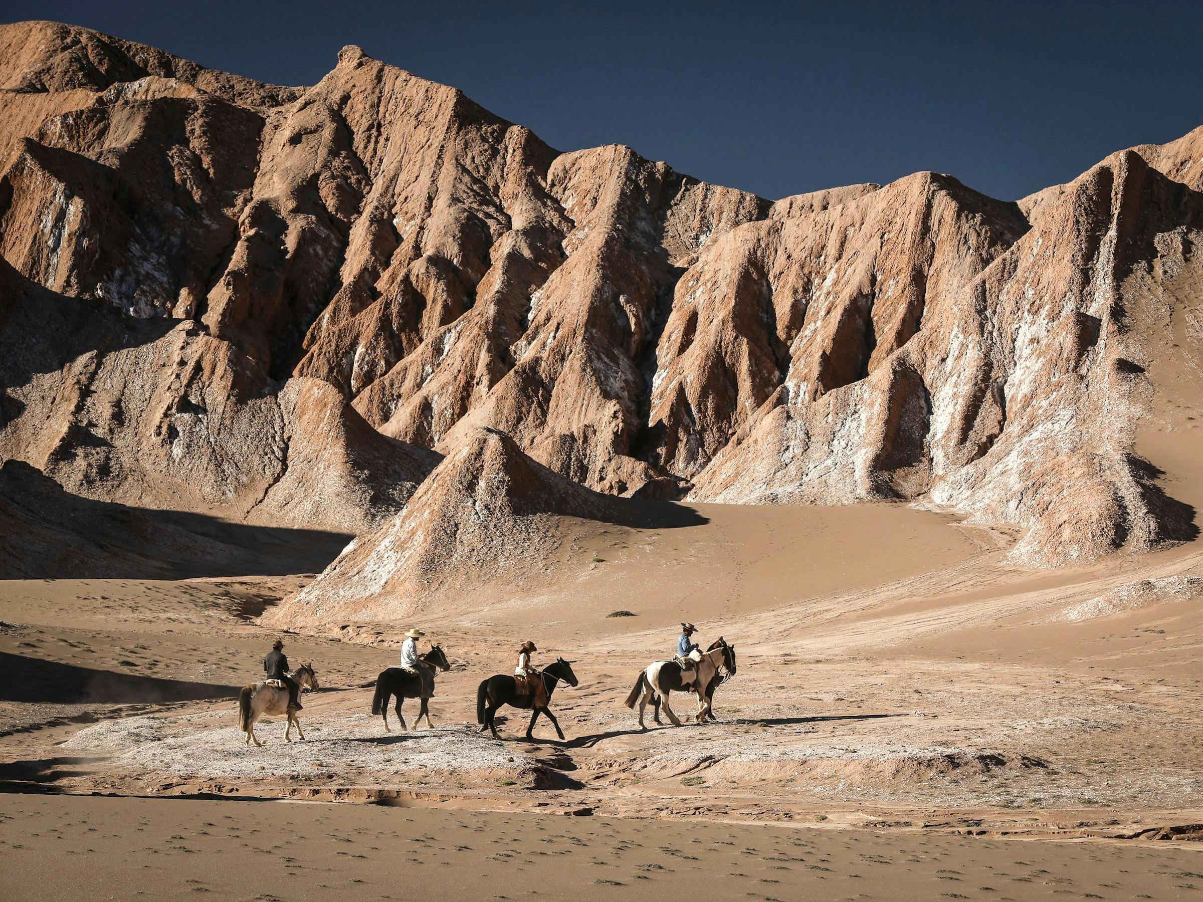 Horseback riders cross a sandy valley beneath tall rocky cliffs.