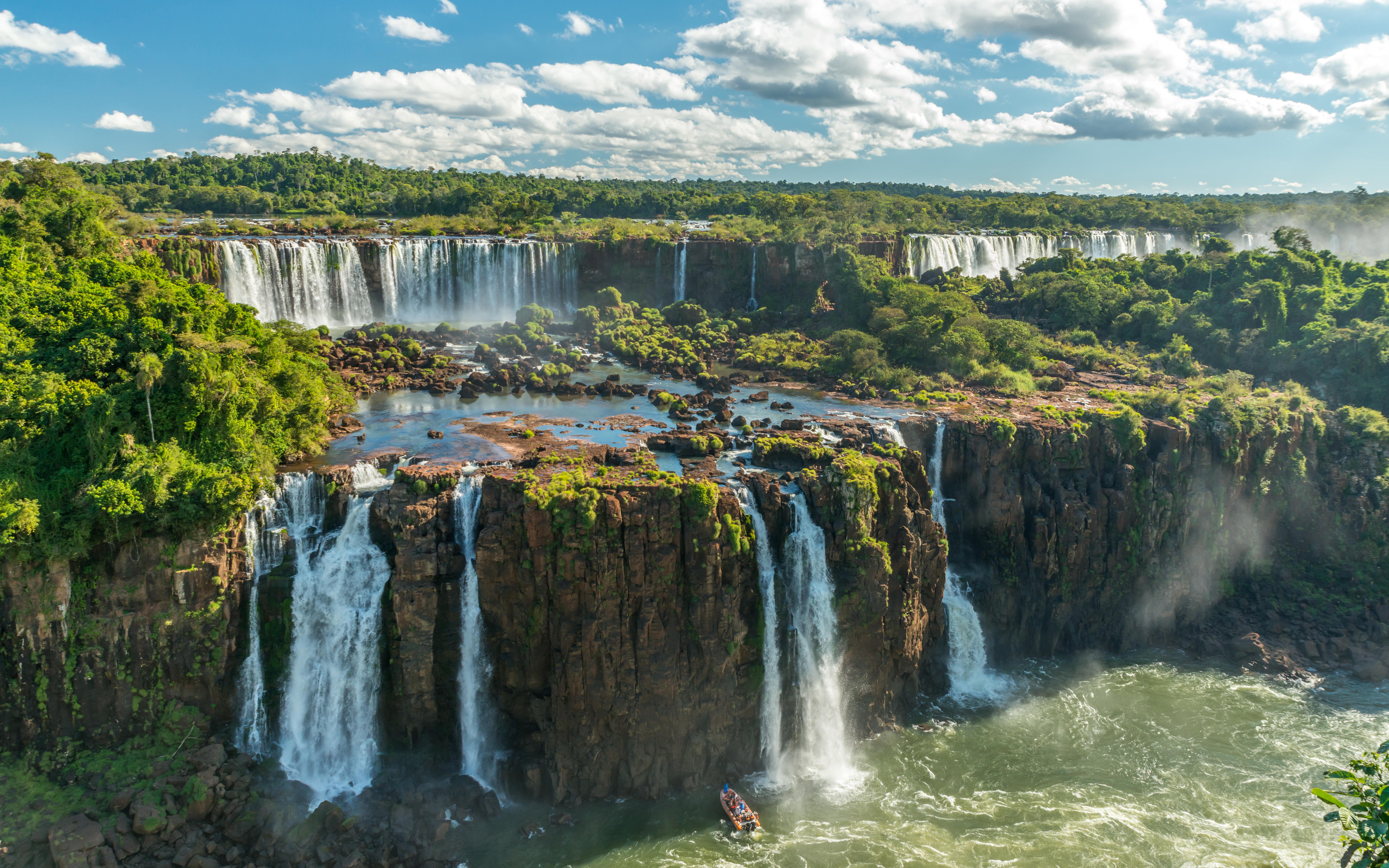 Waterfalls cascade over dark cliffs into swirling river water surrounded by green forest.