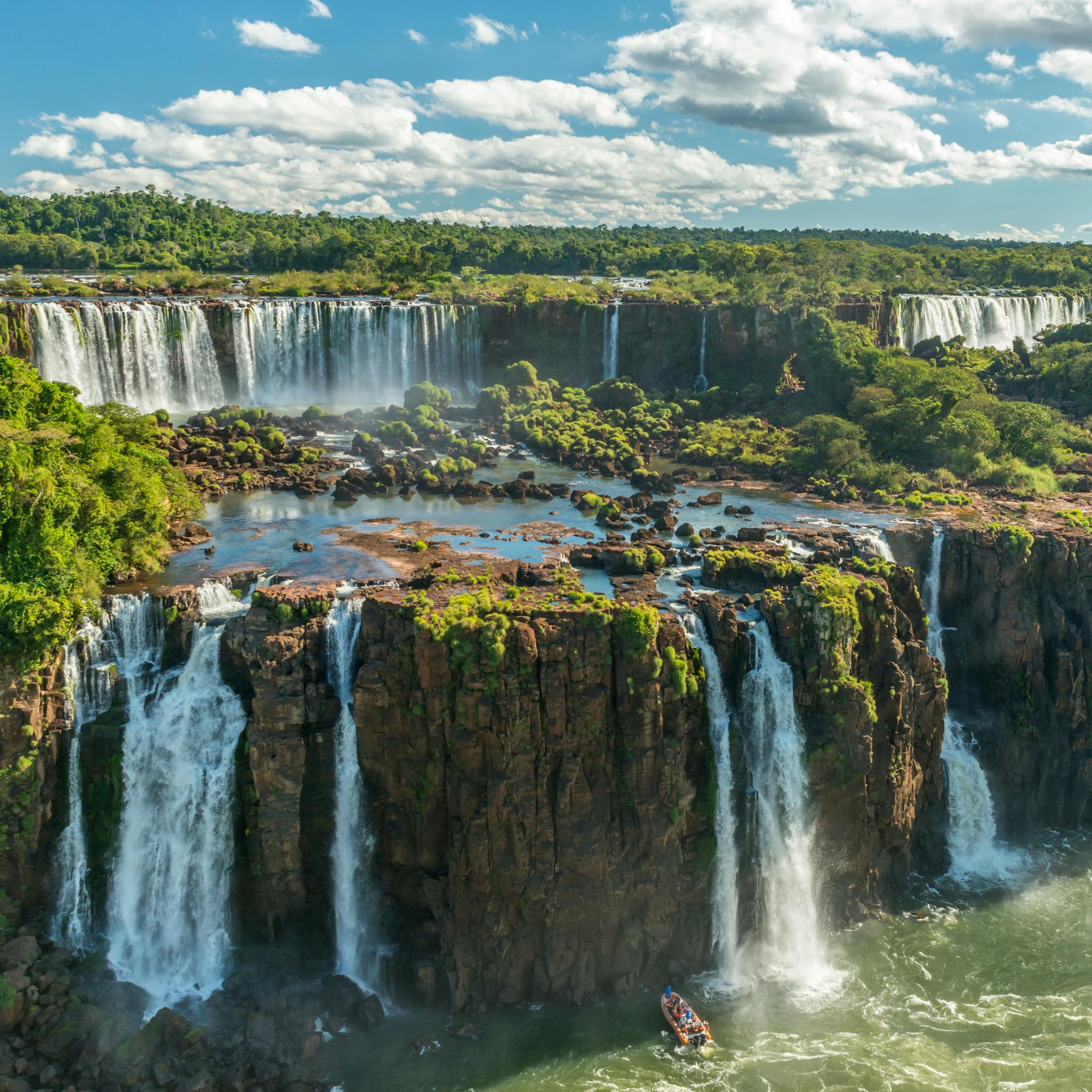 Waterfalls cascade over dark cliffs into swirling river water surrounded by green forest.