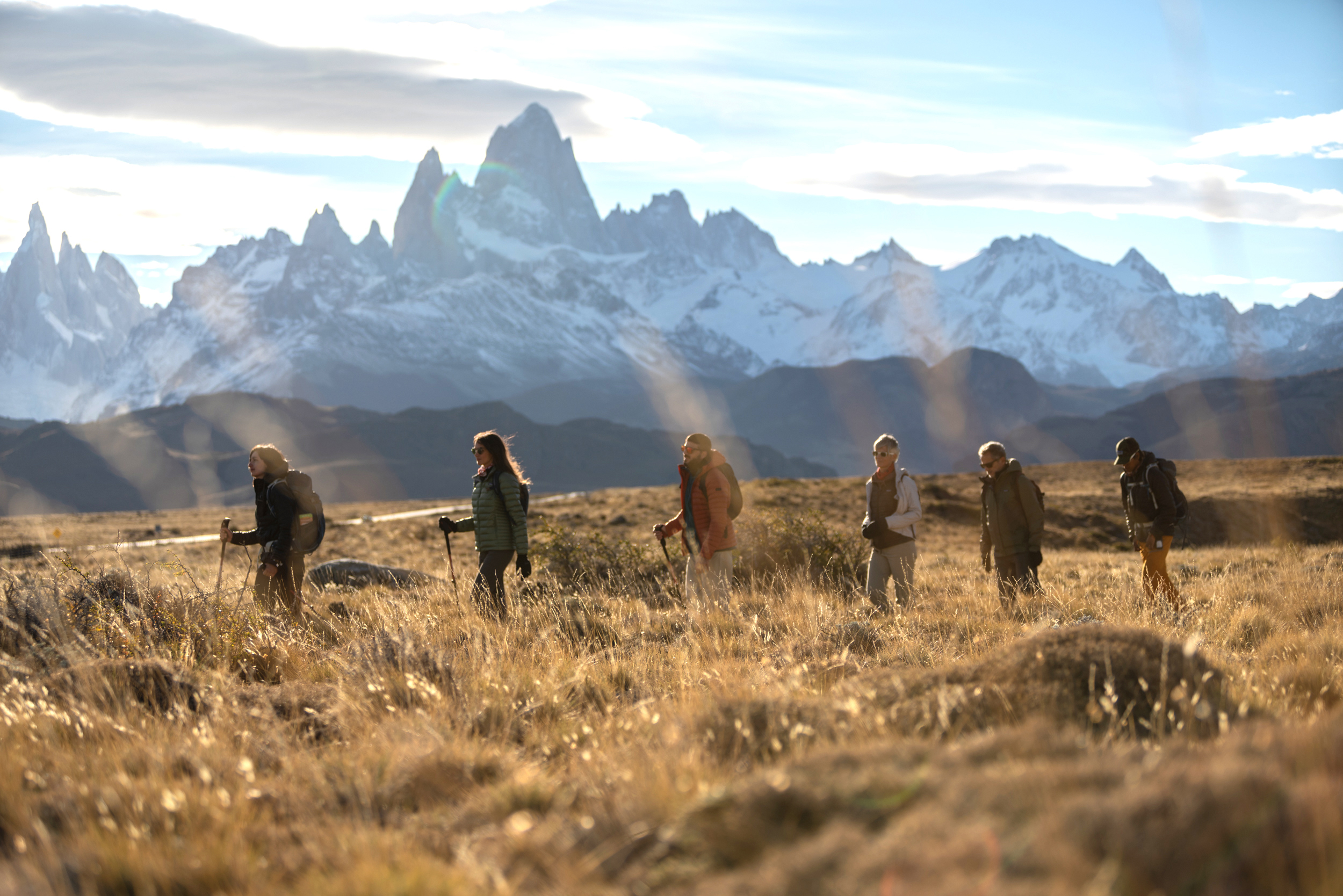 Group of hikers walks through tall grass with jagged mountains in the distance.