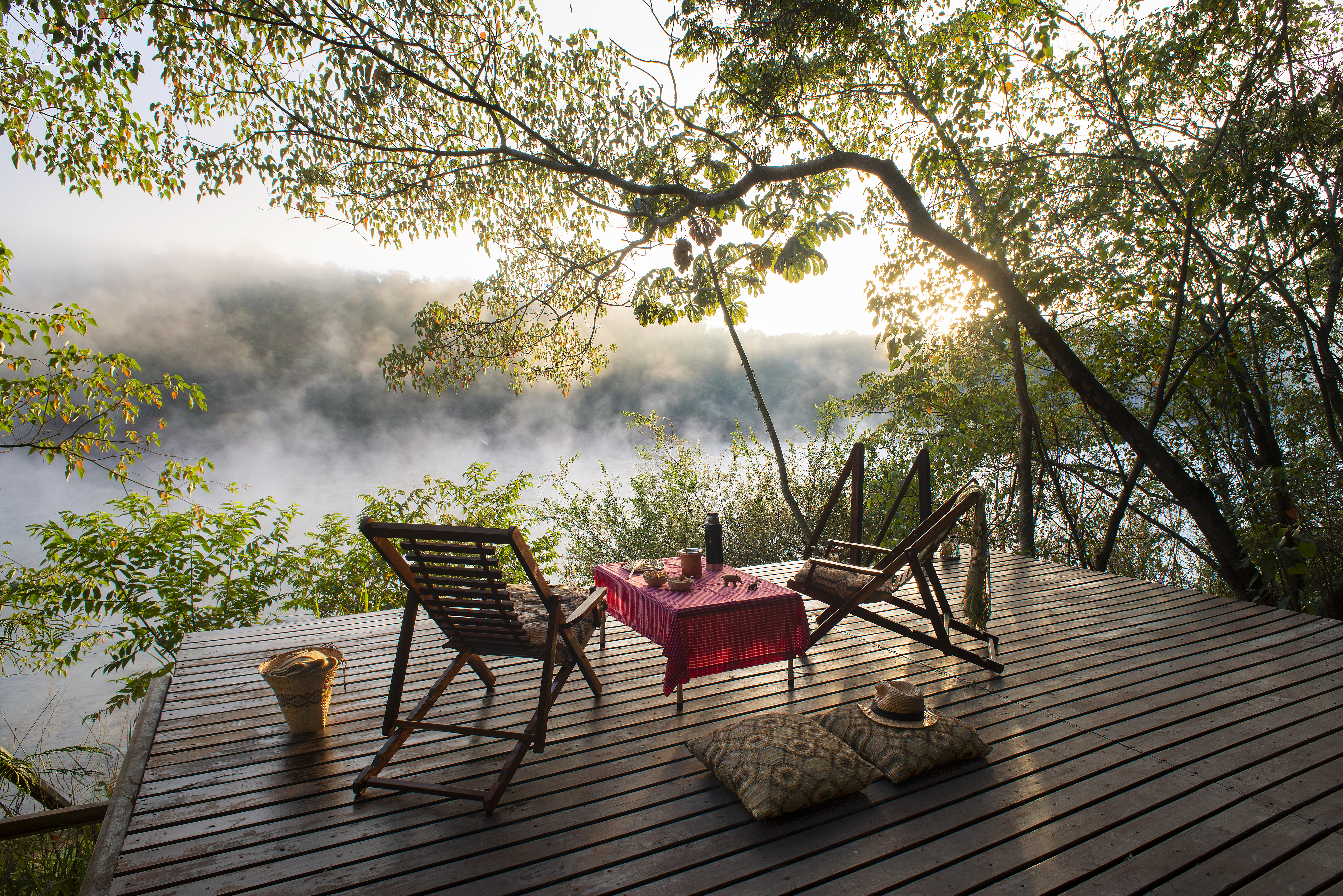 Wooden deck with chairs and a table overlooks misty forest canopy lit by sunrise.