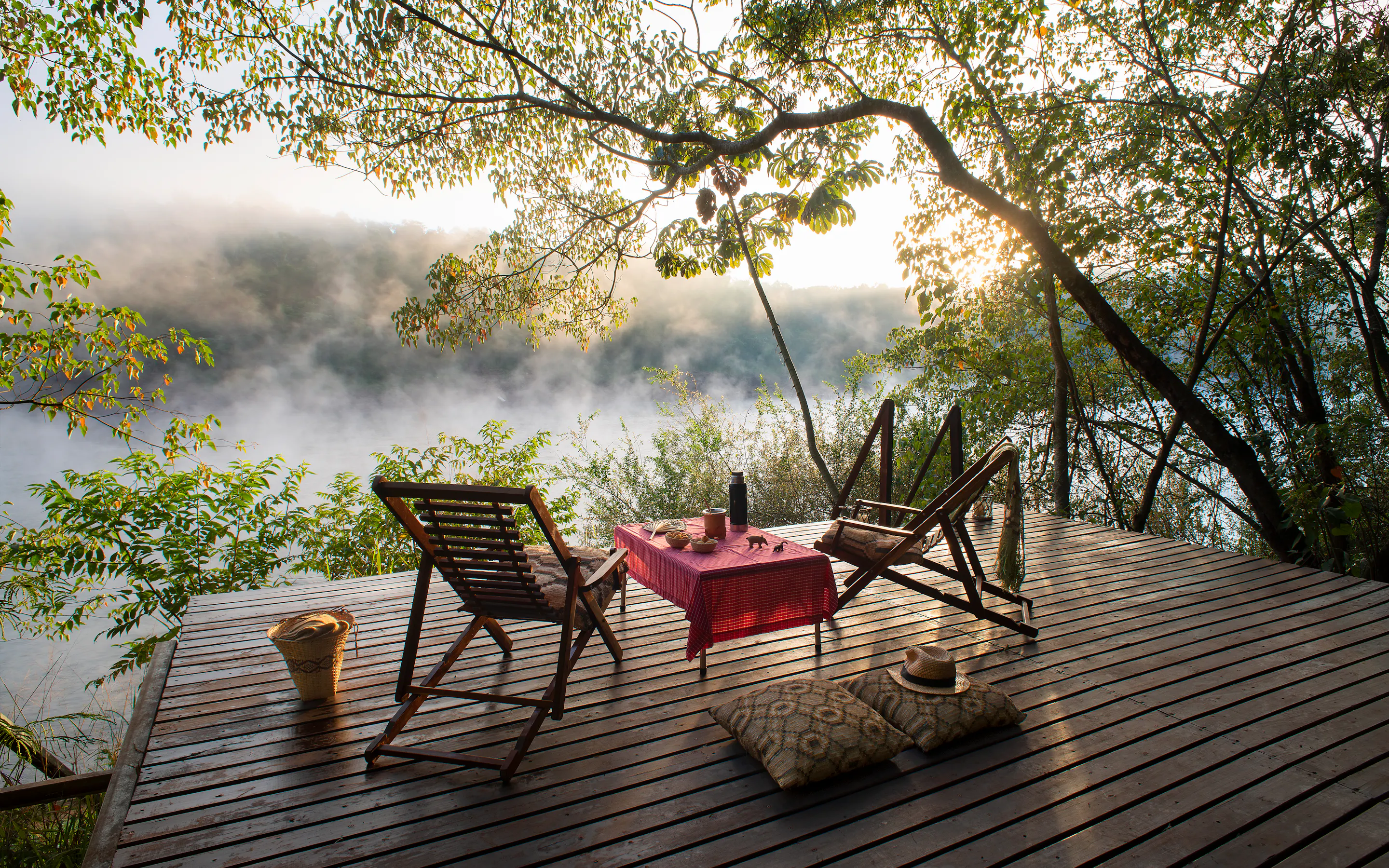 Wooden deck with chairs and a table overlooks misty forest canopy lit by sunrise.