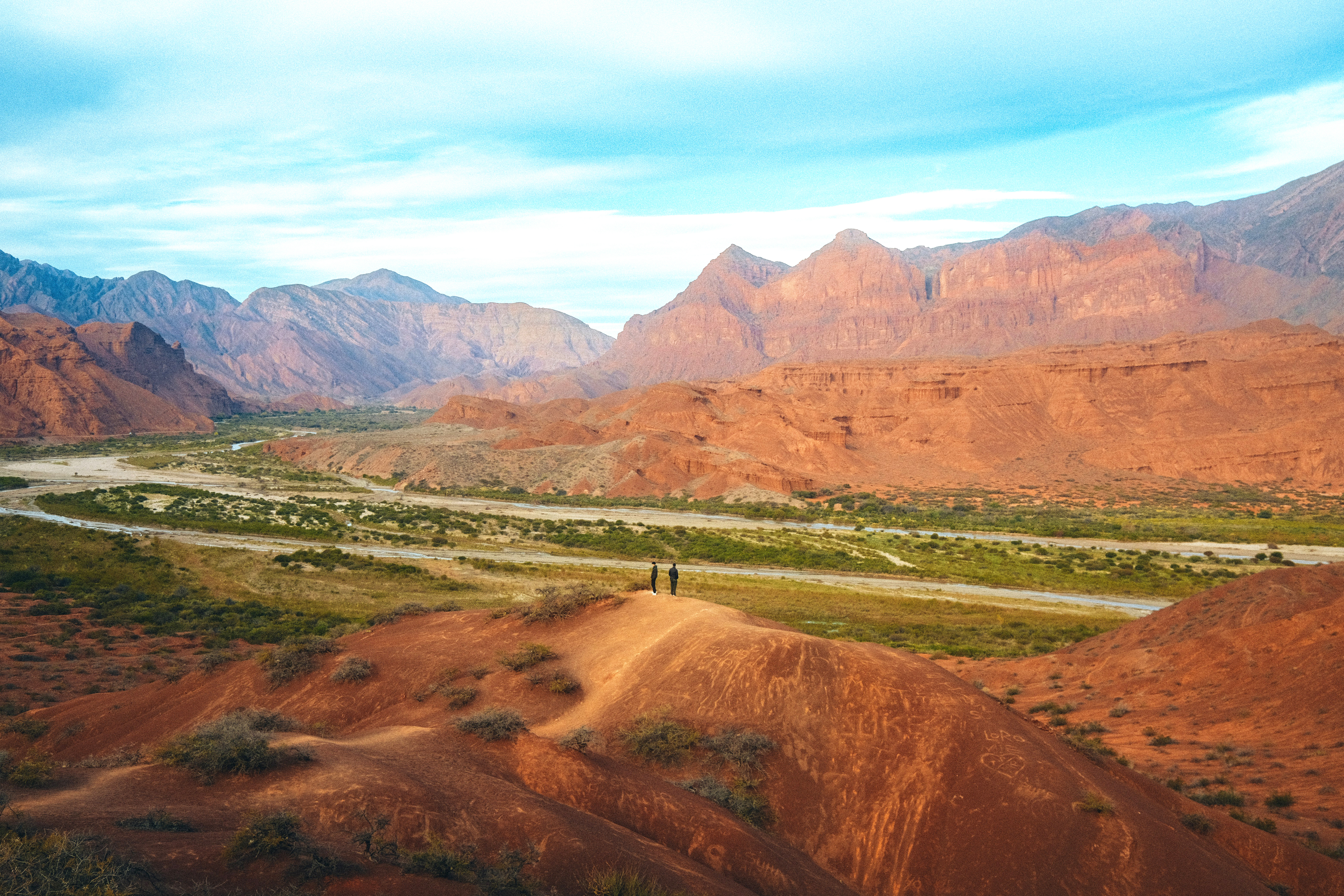 Wide valley with red hills and jagged mountains stretches beneath a pale blue sky.